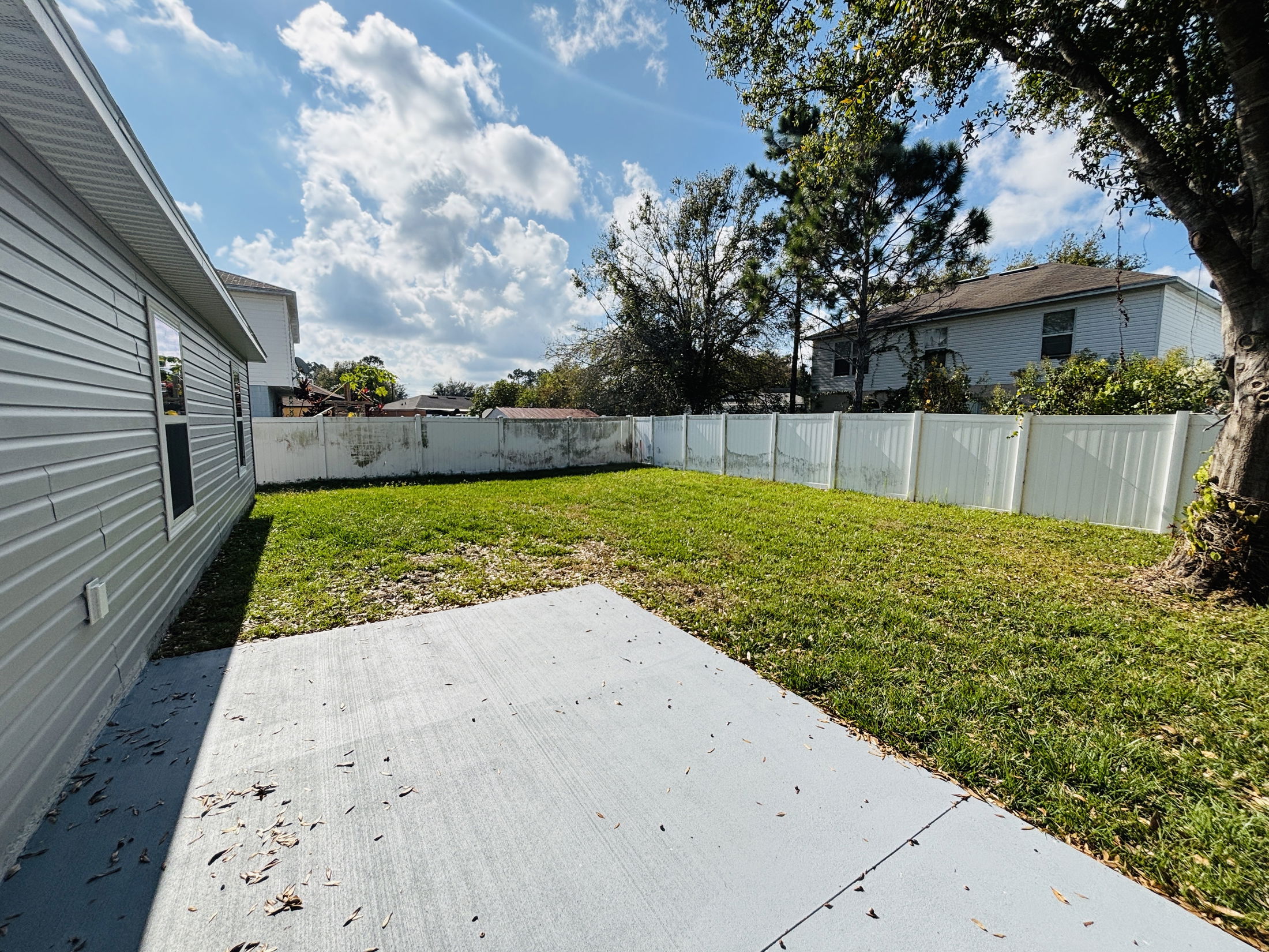 Sunny backyard with a concrete patio, green lawn, and white privacy fence under a blue sky.