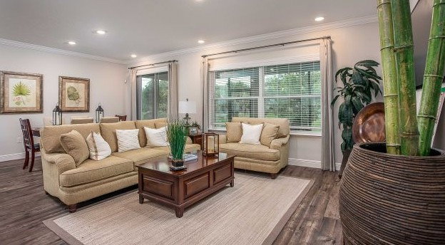 Spacious modern living room with a beige sofa, decorative pillows, wooden coffee table, and large windows surrounded by greenery.