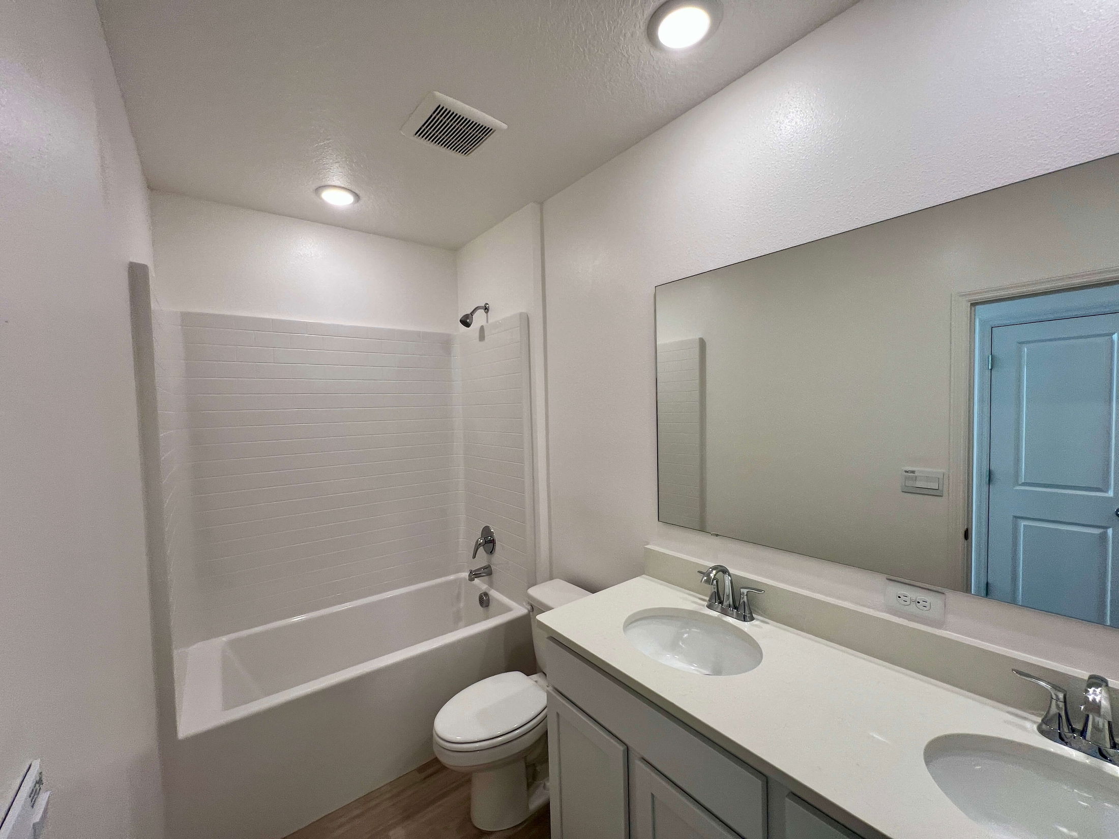 Modern bathroom with a white bathtub and shower, dual sinks, large mirror, toilet, and bright overhead lighting.