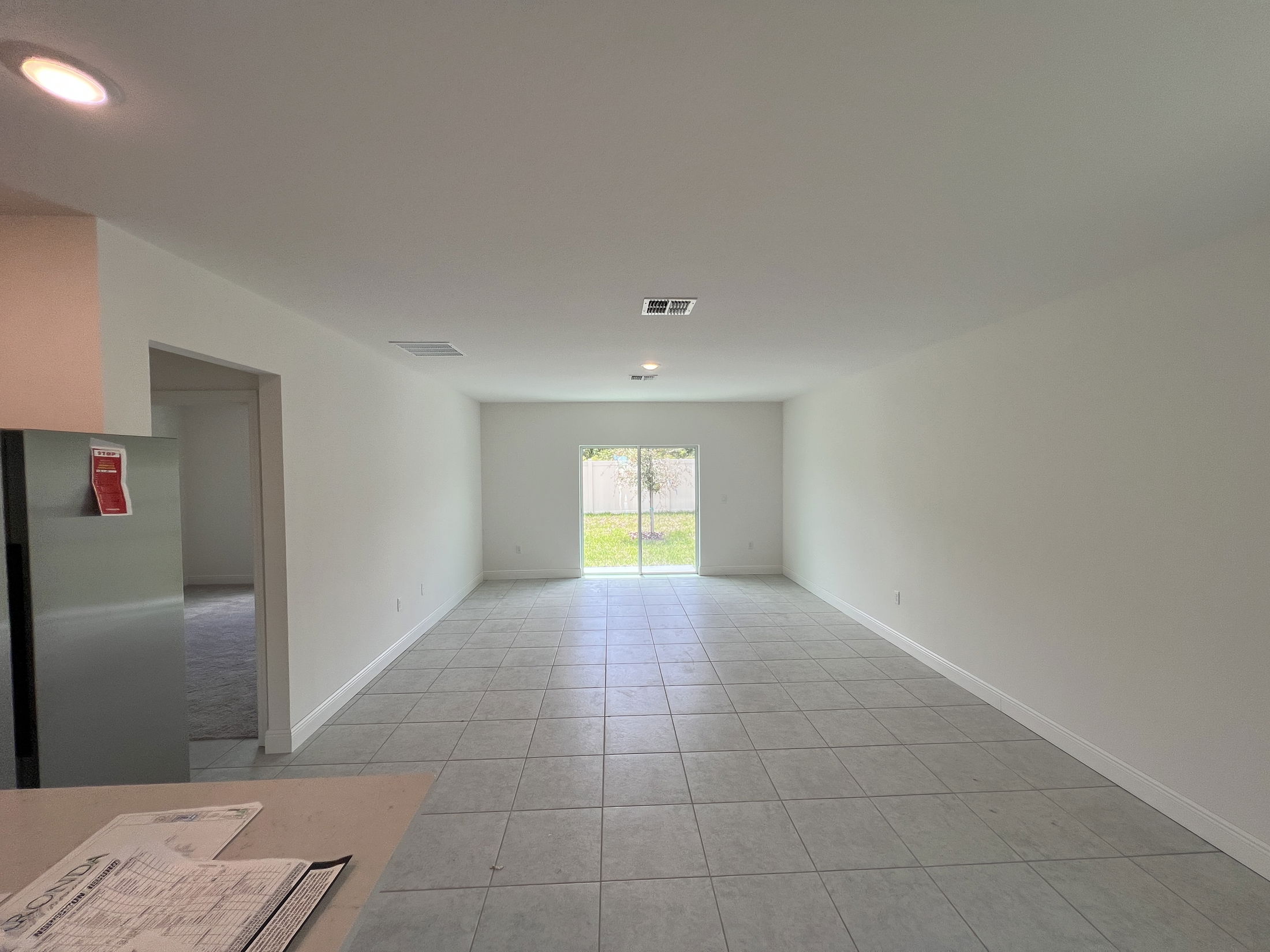 Spacious, unfurnished living room with tile flooring, neutral walls, and a sliding glass door leading to a fenced backyard.