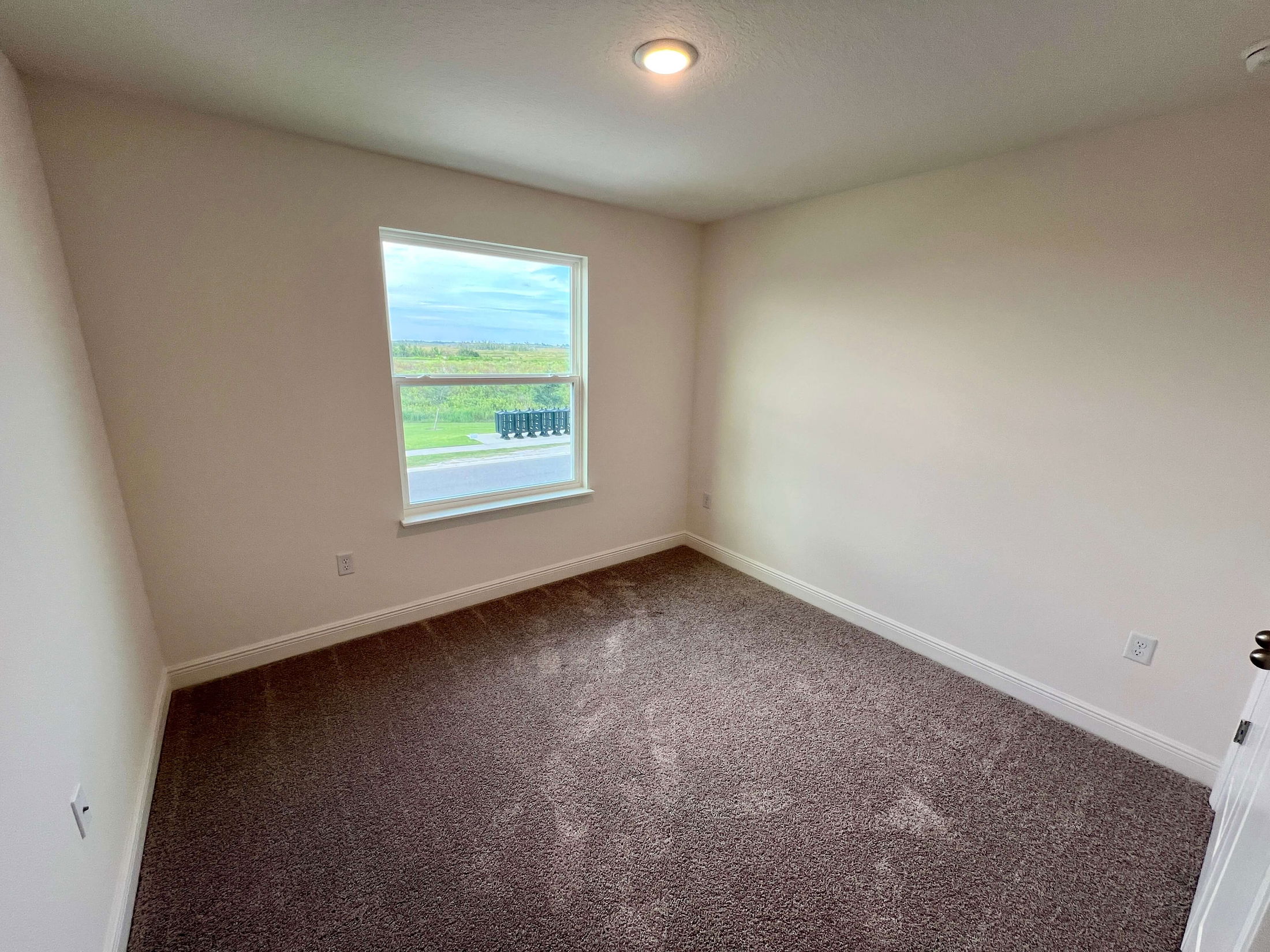 Bright, empty bedroom with beige carpet and a large window overlooking a green outdoor area.