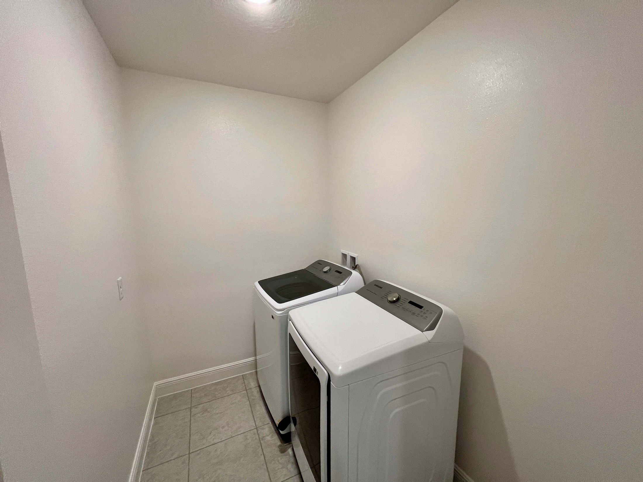 A small laundry room features a modern top-loading washer and dryer set against plain white walls and tiled flooring.