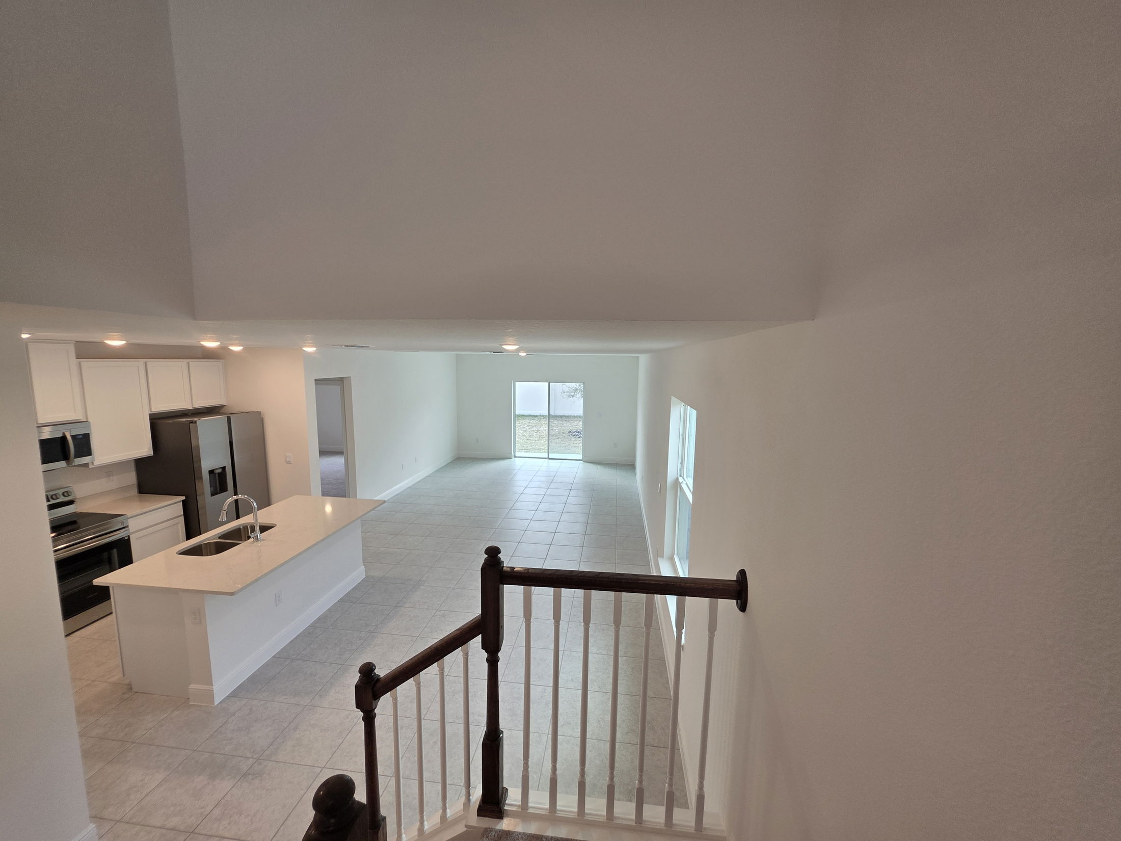 A spacious, modern living area with an open kitchen layout, featuring white cabinets and stainless steel appliances, viewed from an upper staircase.
