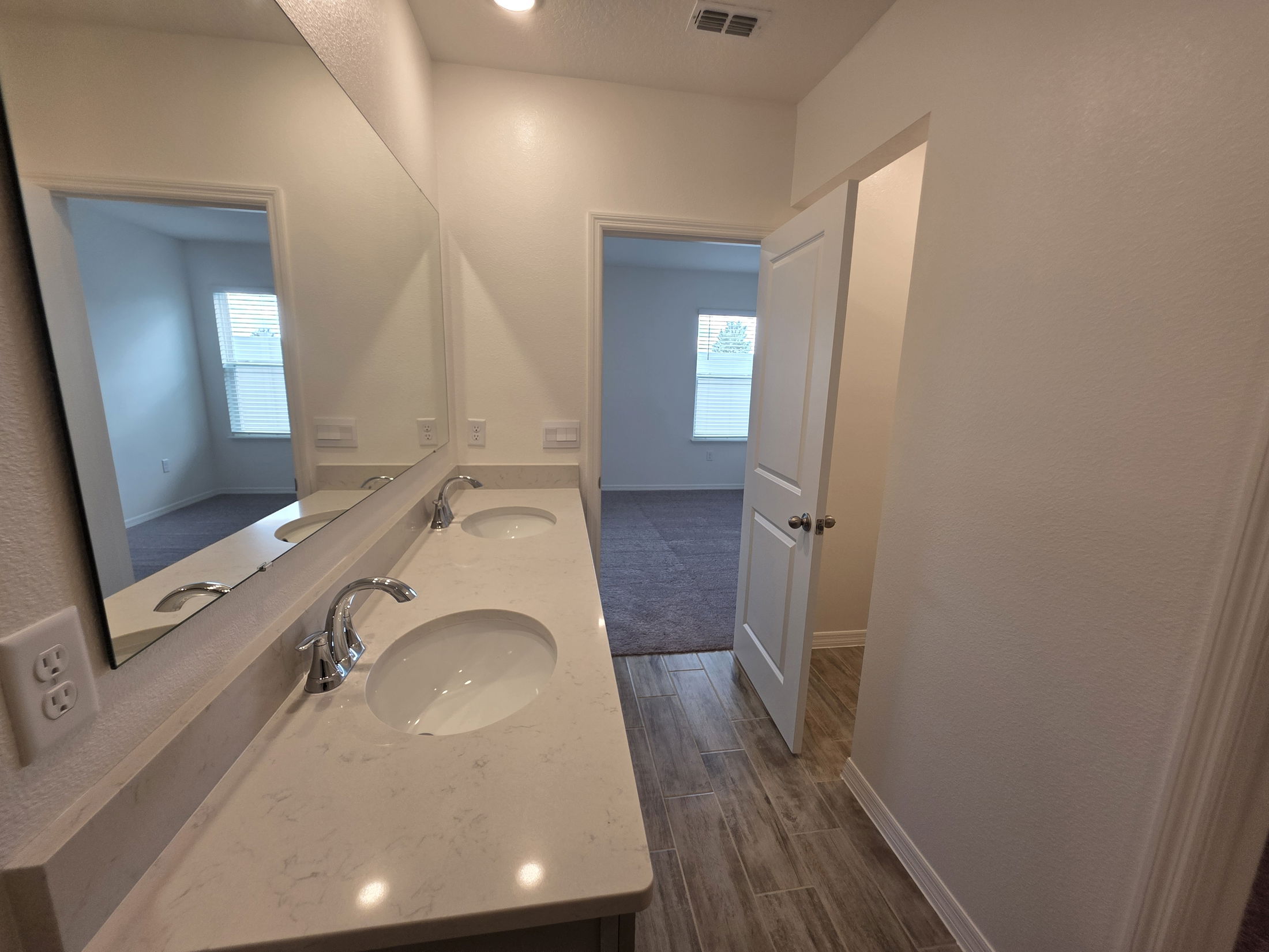 Modern bathroom interior featuring a dual sink vanity, large mirror, and doorway leading to a carpeted room with natural light.