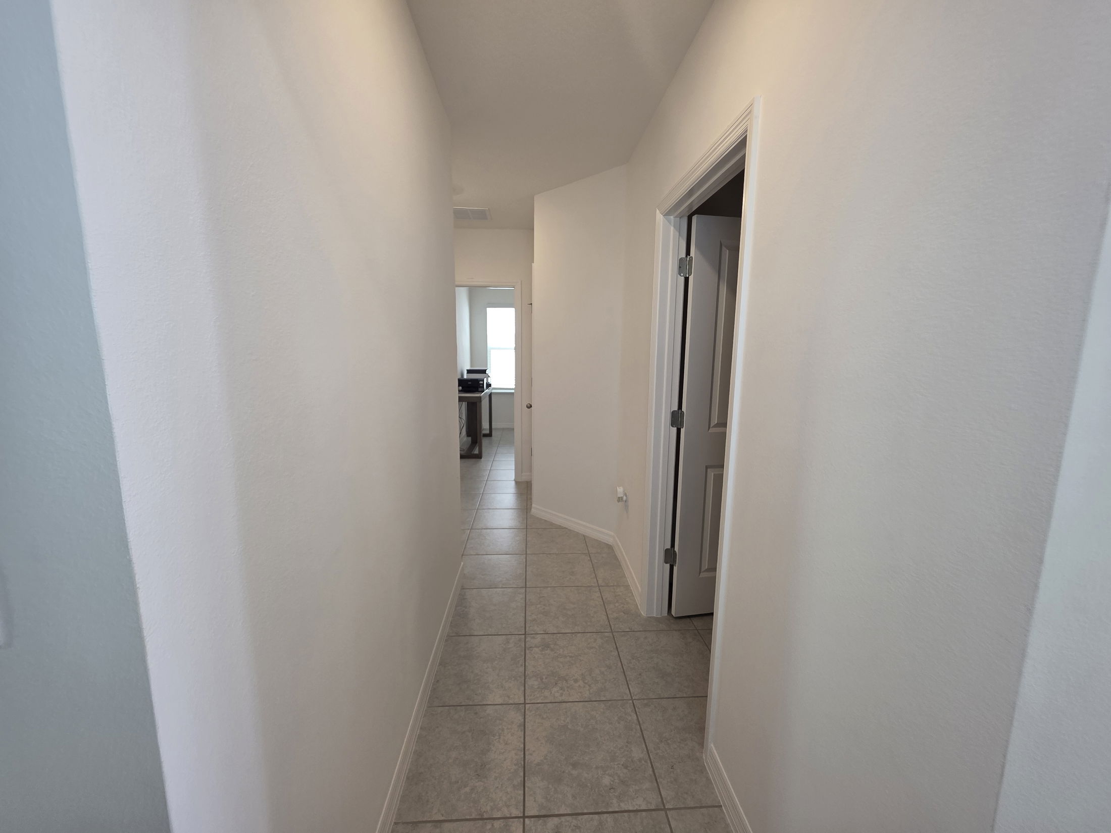 A well-lit hallway in a modern home featuring white walls and tile flooring leading to an open area.