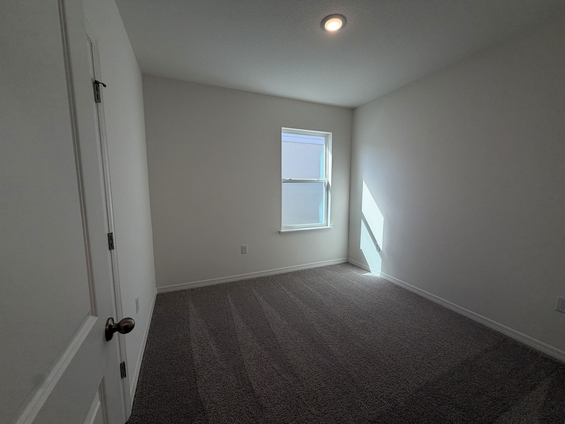 A bright and empty bedroom featuring neutral walls, carpeted flooring, and a single window letting in natural light.