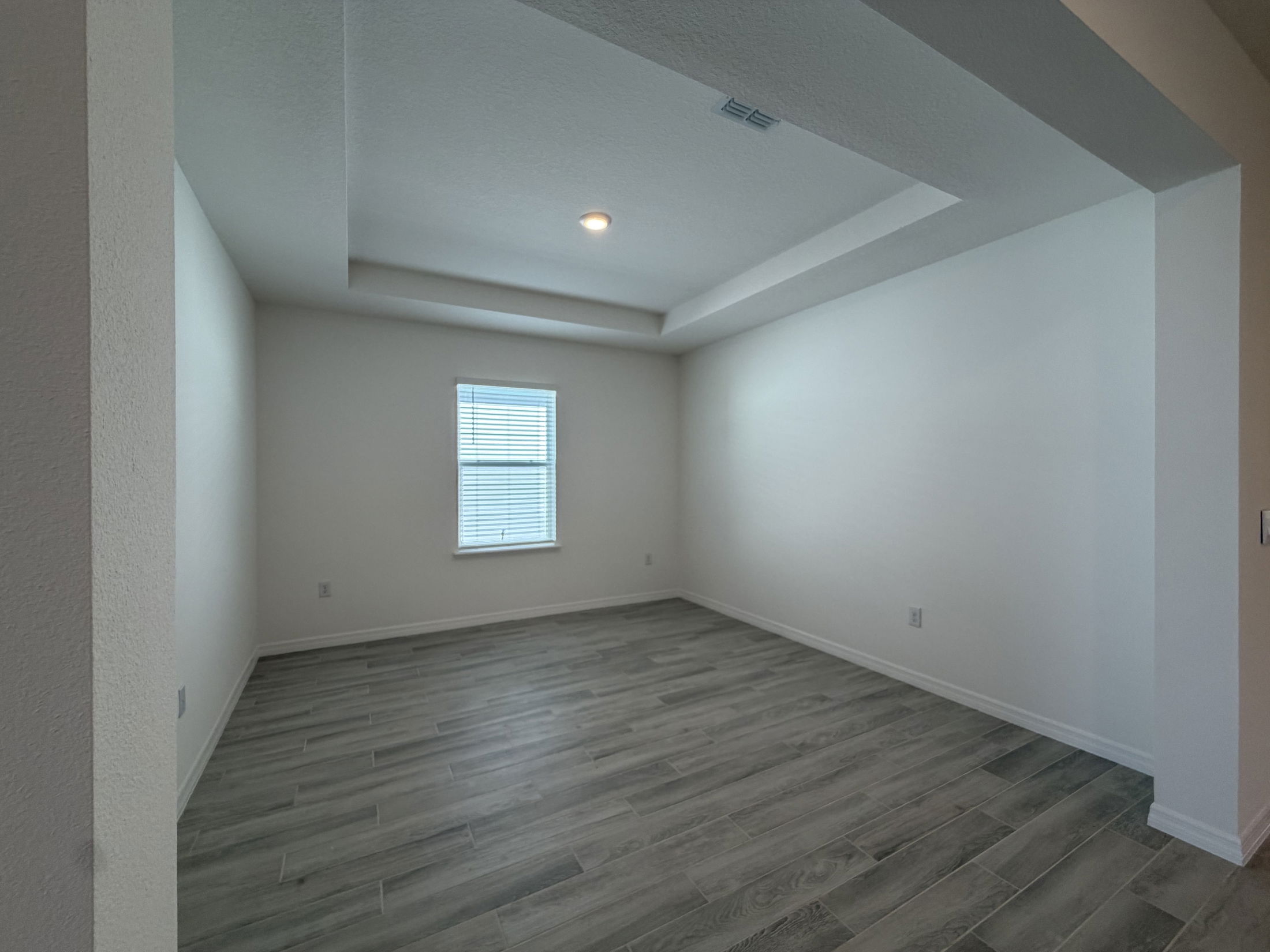Empty modern living room with gray hardwood flooring and a window featuring blinds.