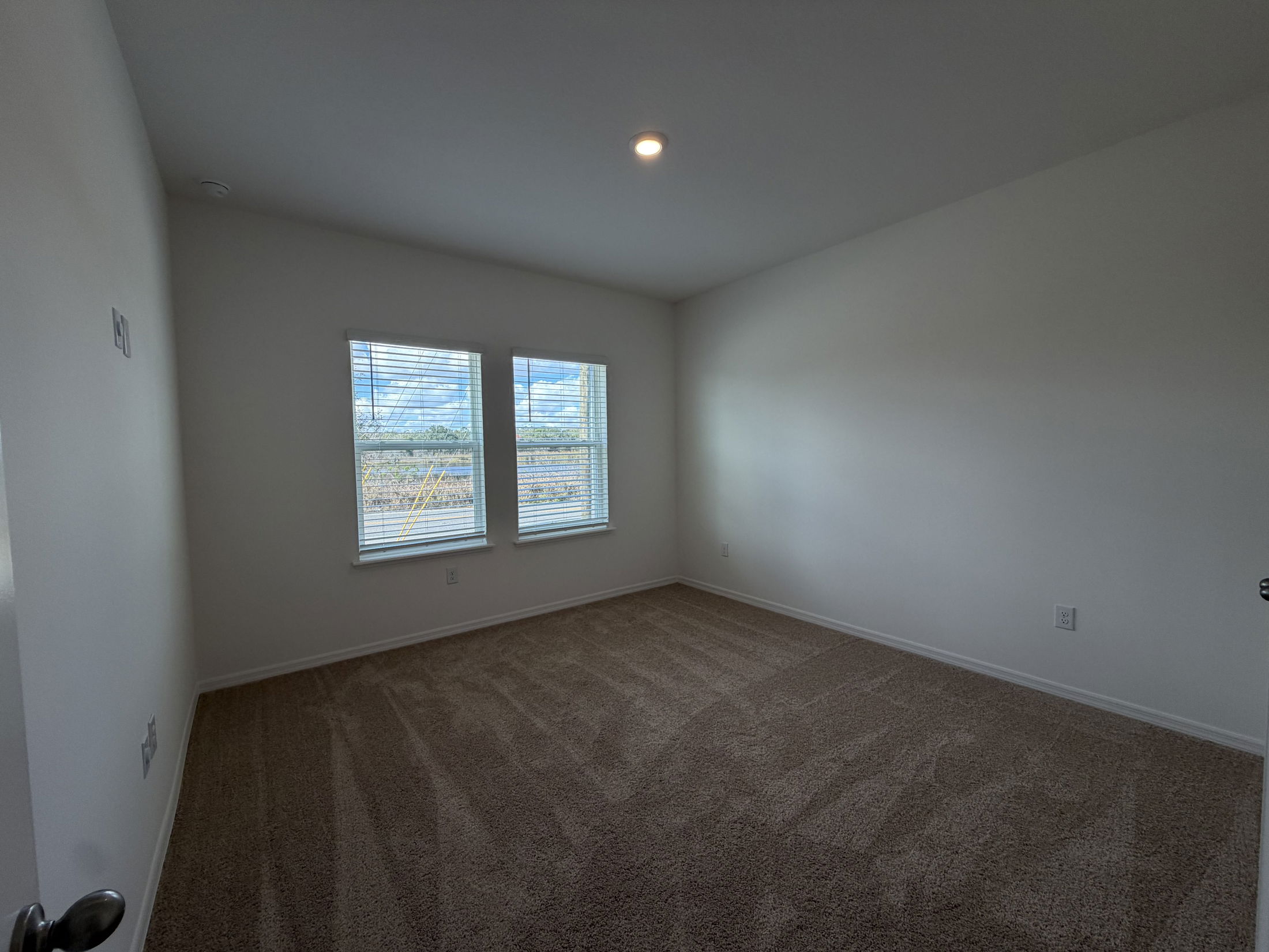 A bright and spacious empty bedroom featuring two windows with blinds, neutral-colored walls, and carpeted flooring.