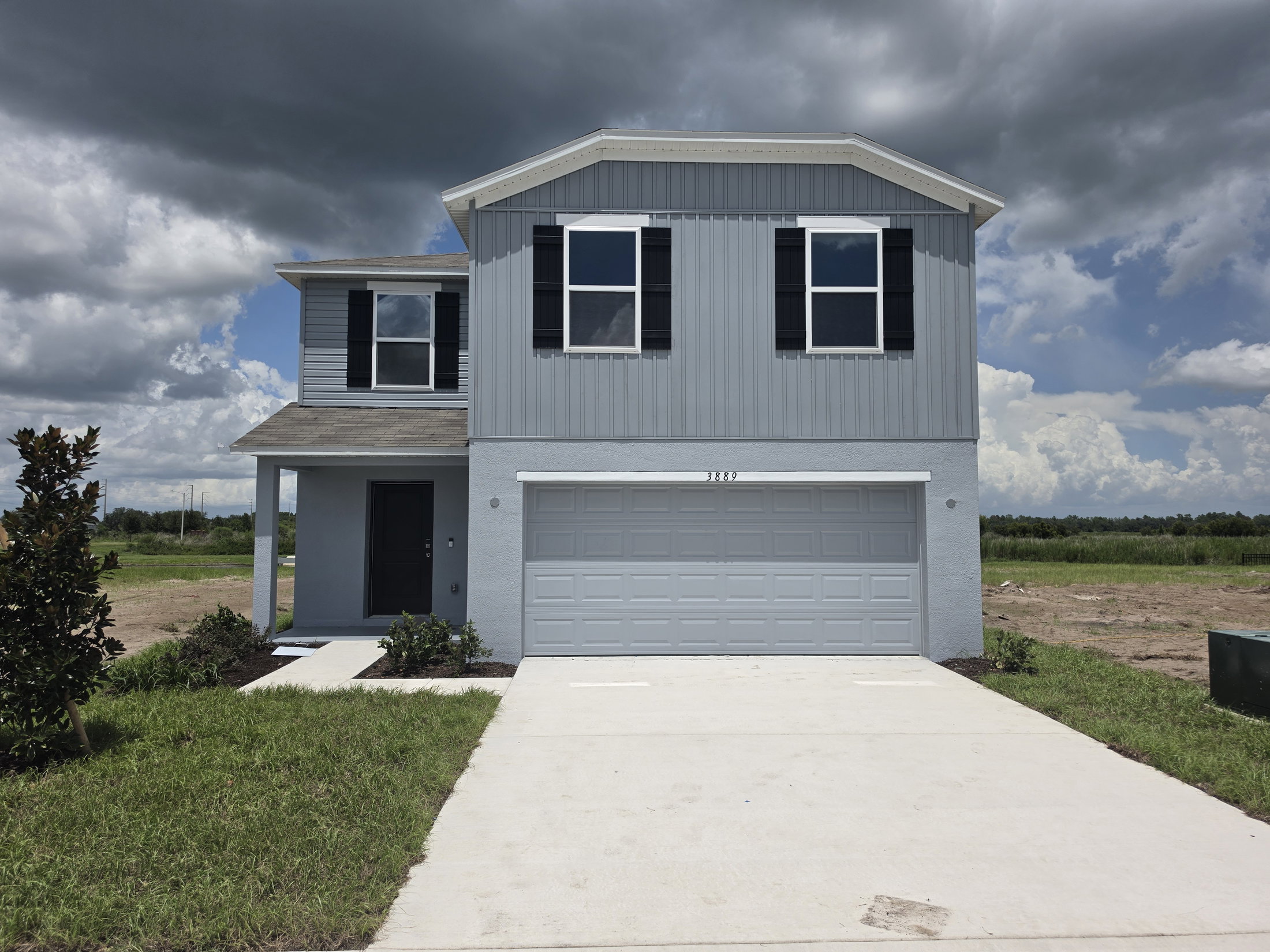 A two-story blue house with black shutters and a gray garage door, surrounded by grass and under a cloudy sky.