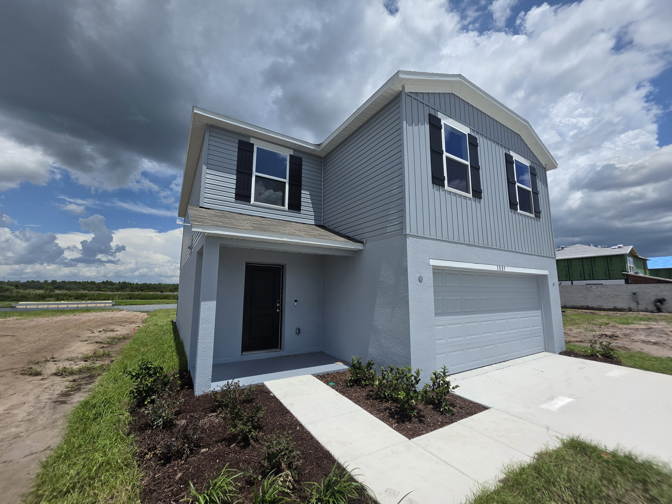 A two-story gray house with black shutters and a garage, surrounded by freshly planted landscaping and a cloudy sky.