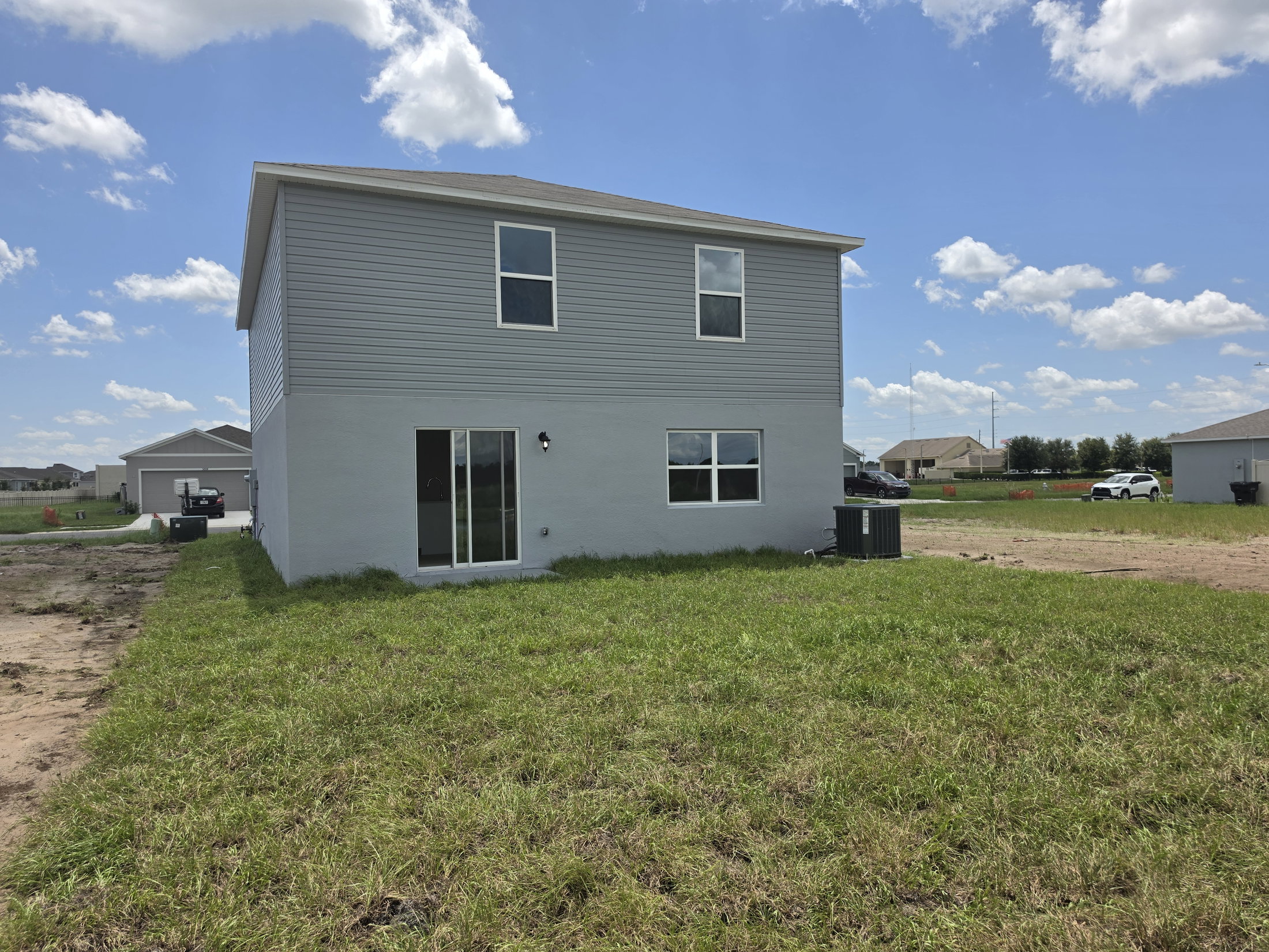 A two-story gray house with a sliding glass door and several windows, surrounded by a grassy area under a blue sky with scattered clouds.