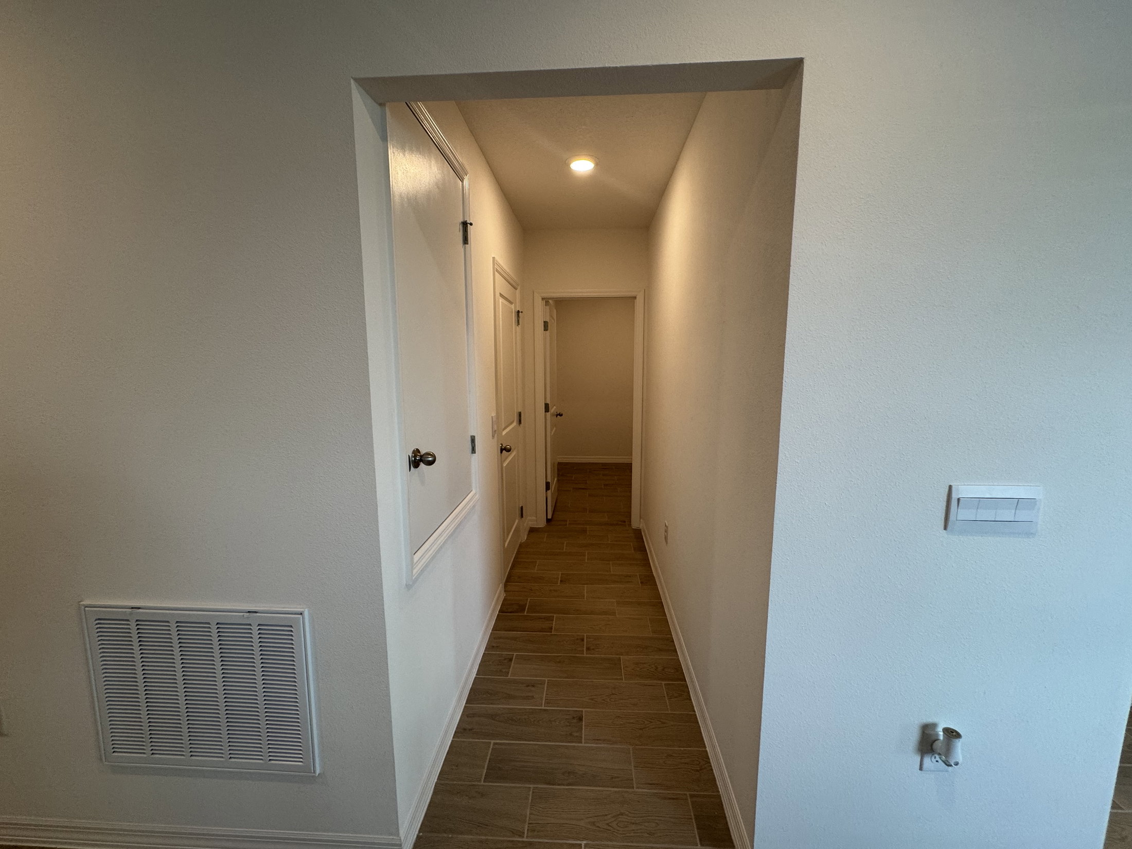Narrow hallway with beige tile flooring and white walls, featuring a door on the left and another doorway at the end.
