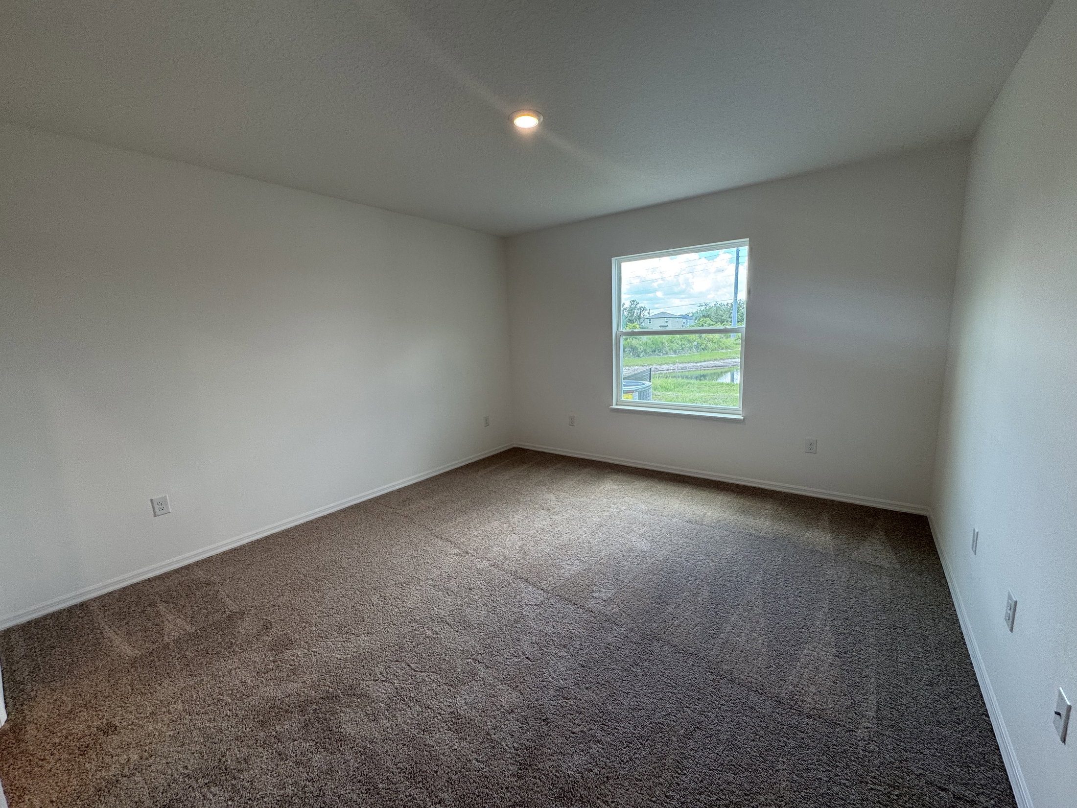 Empty, well-lit room with beige carpeting and a window overlooking a green landscape.