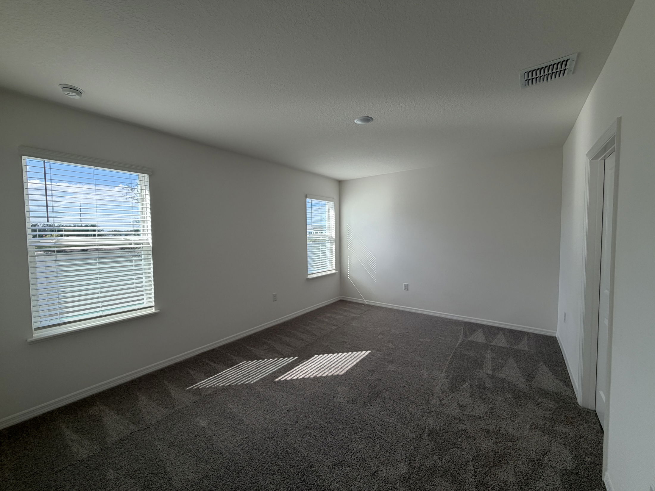 Spacious empty room with gray carpet, white walls, and two windows with blinds letting in natural light.