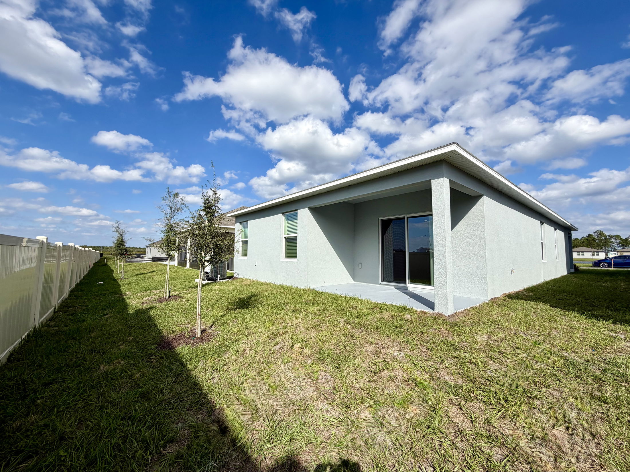 Backyard view of a modern house with sliding glass door, partially enclosed patio, and young trees along a white fence under a blue sky.
