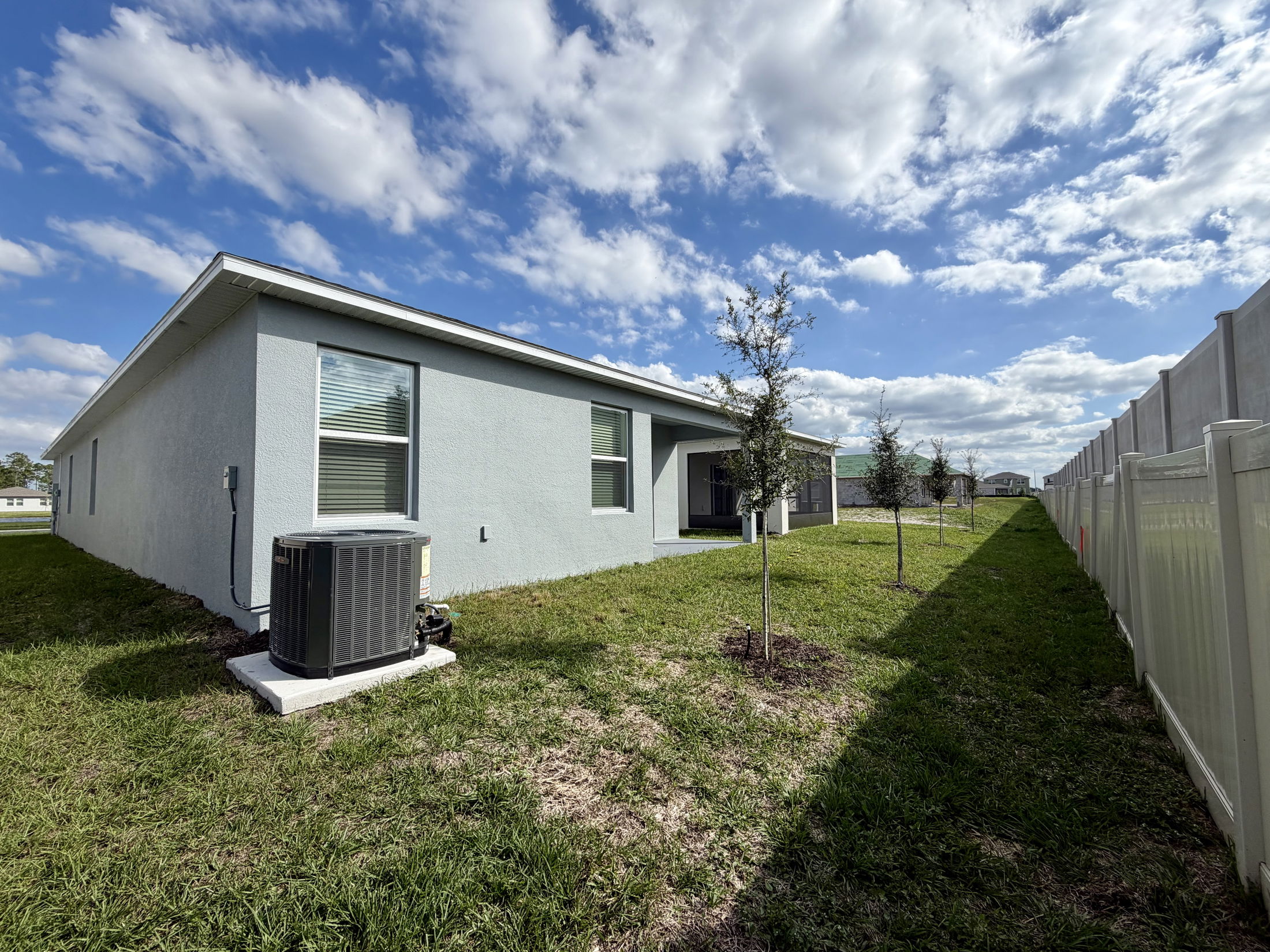 Side view of a suburban single-story house with an HVAC unit on a grassy lawn, set against a blue sky with scattered clouds.