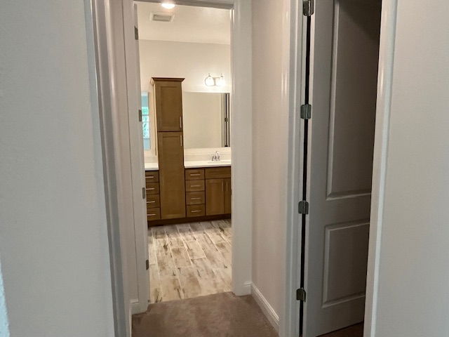 Narrow hallway with beige walls leading to a modern bathroom featuring wooden cabinets and a large mirror.
