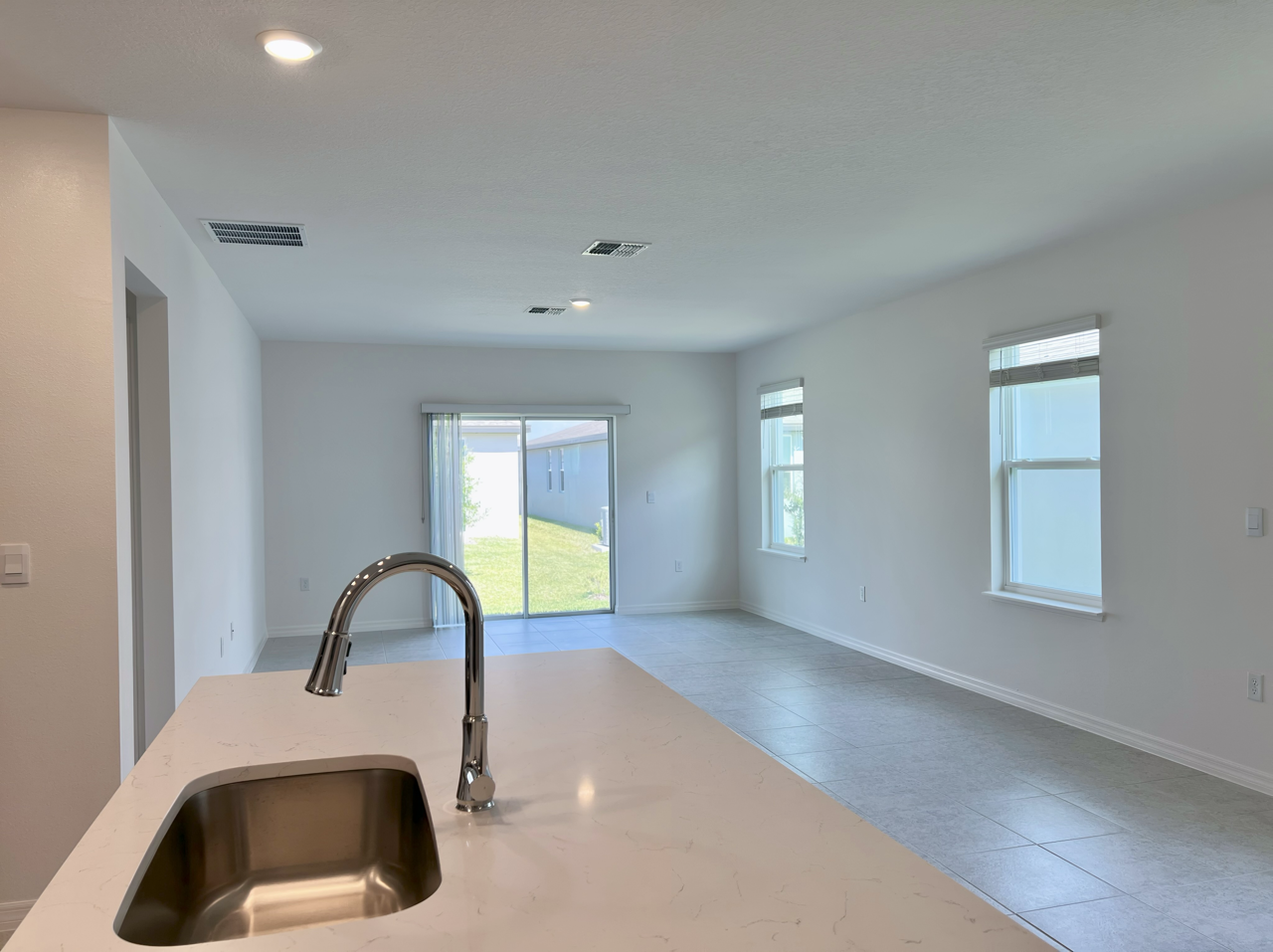 Modern, spacious open-plan living area with large windows and a kitchen island, featuring a sleek faucet and a view of the backyard through sliding glass doors.