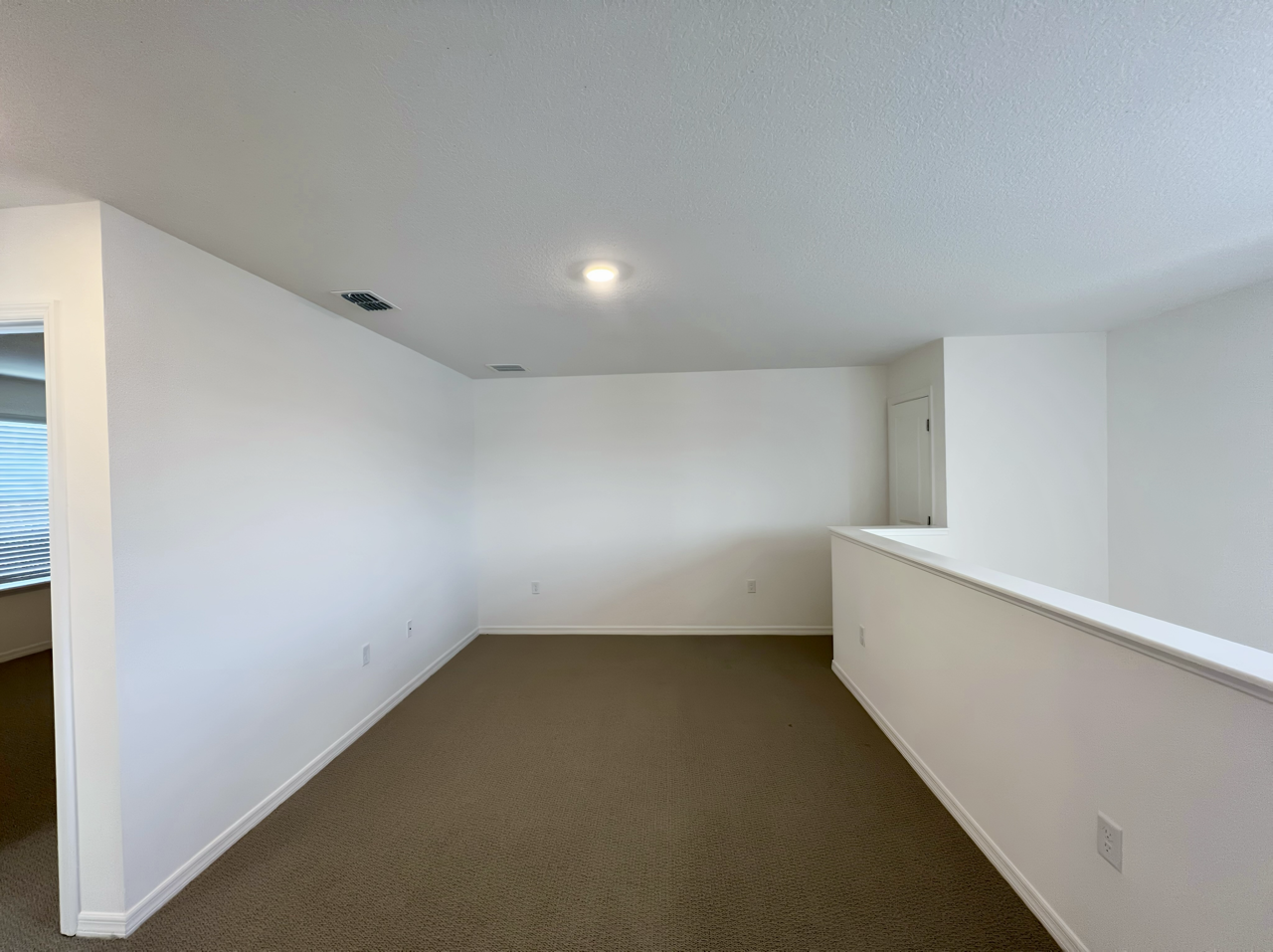 Empty white room with beige carpet and a single ceiling light, featuring a small wall partition on the right.