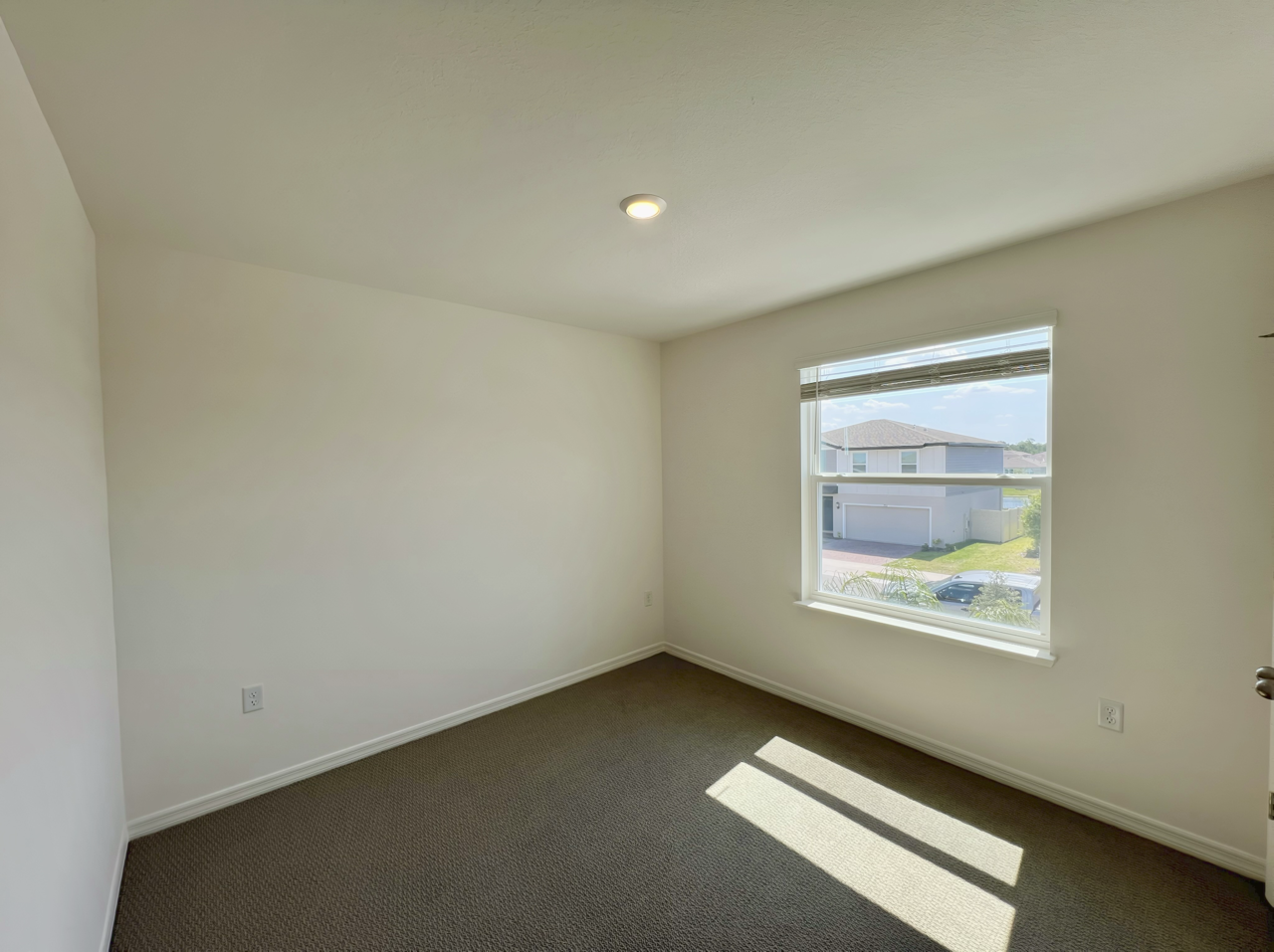 Bright, empty room with beige carpet and a sunny window overlooking a suburban neighborhood.