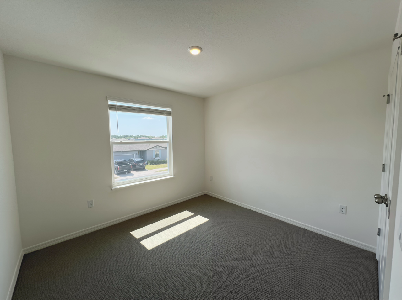 Empty room with white walls, carpeted floor, a single window providing natural light, and a view of houses and cars outside.