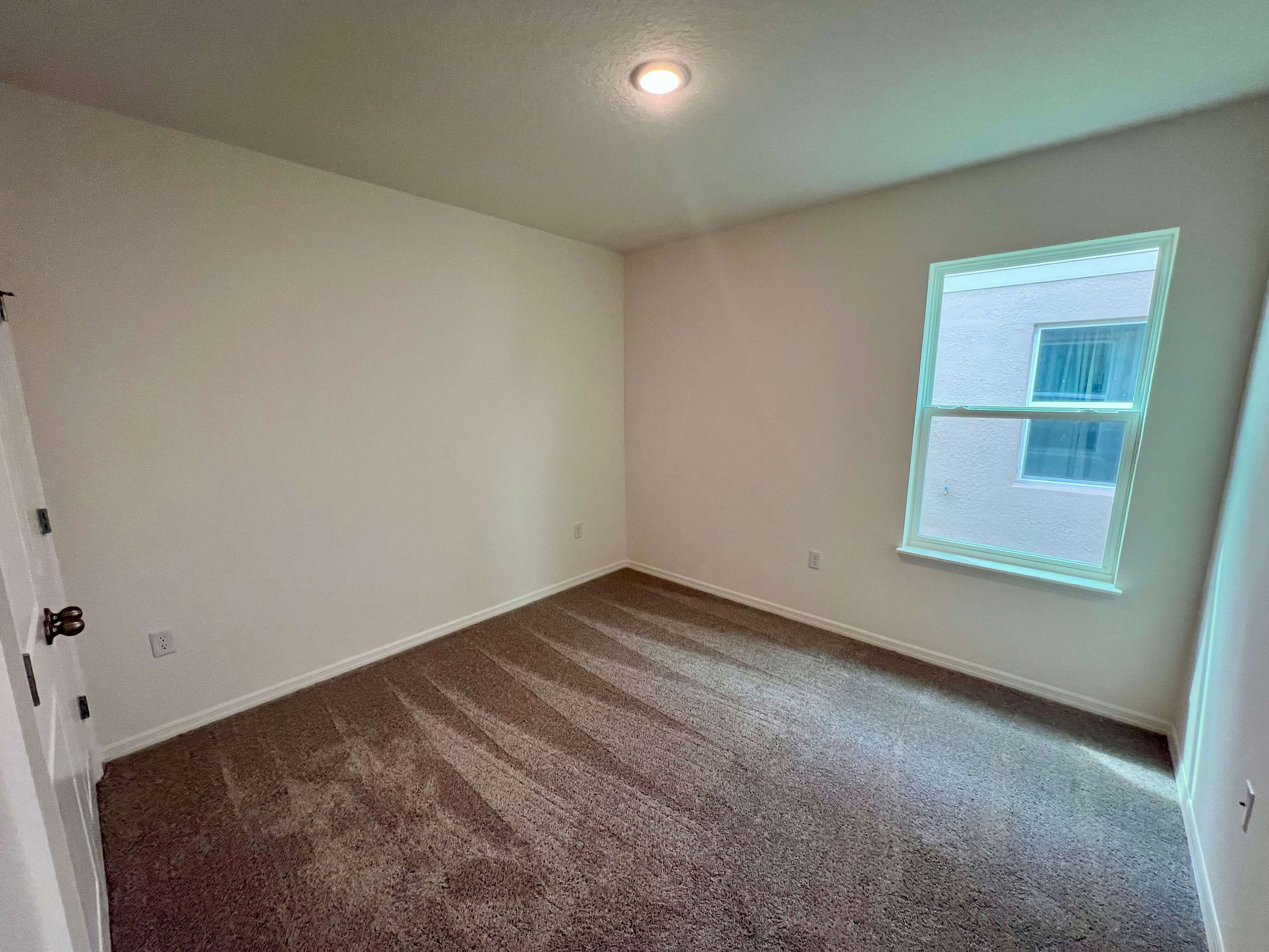 Empty room with beige carpet, a single window, and white walls illuminated by a ceiling light.