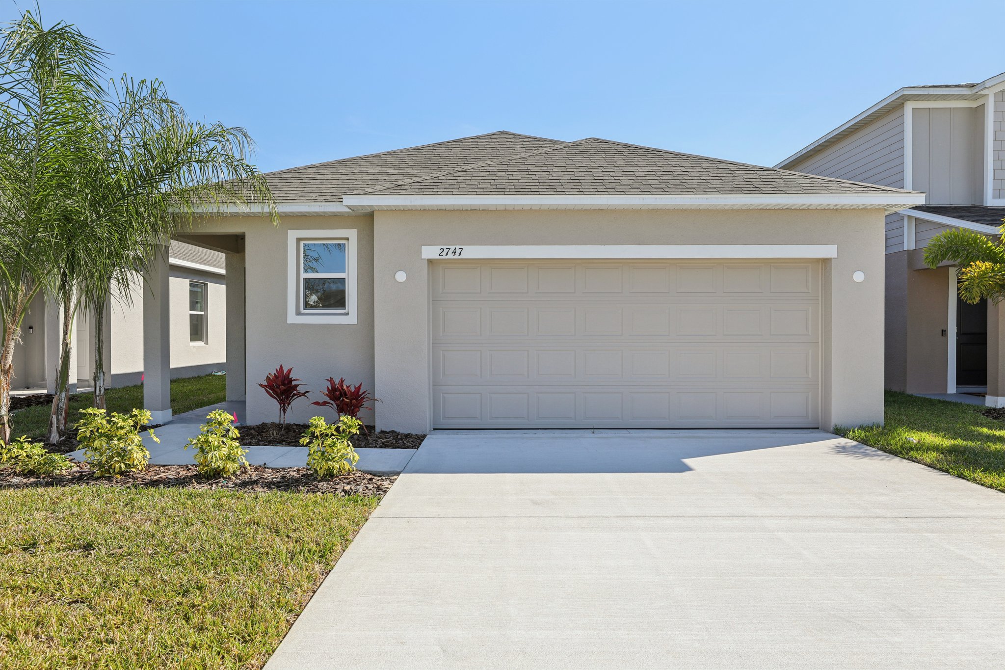 Modern single-story house with a two-car garage, tropical landscaping, and a well-maintained lawn under a clear blue sky.