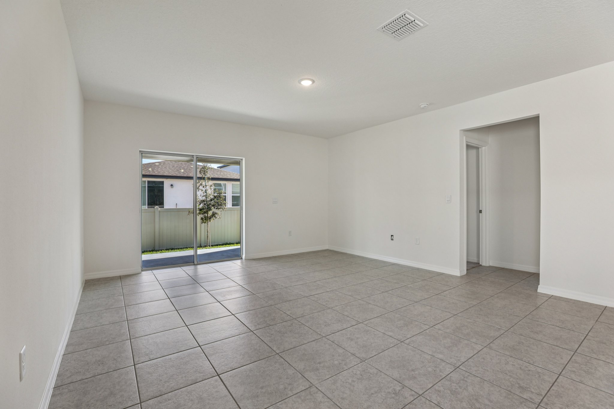 Empty modern living room with tile flooring, white walls, and sliding glass door to backyard.