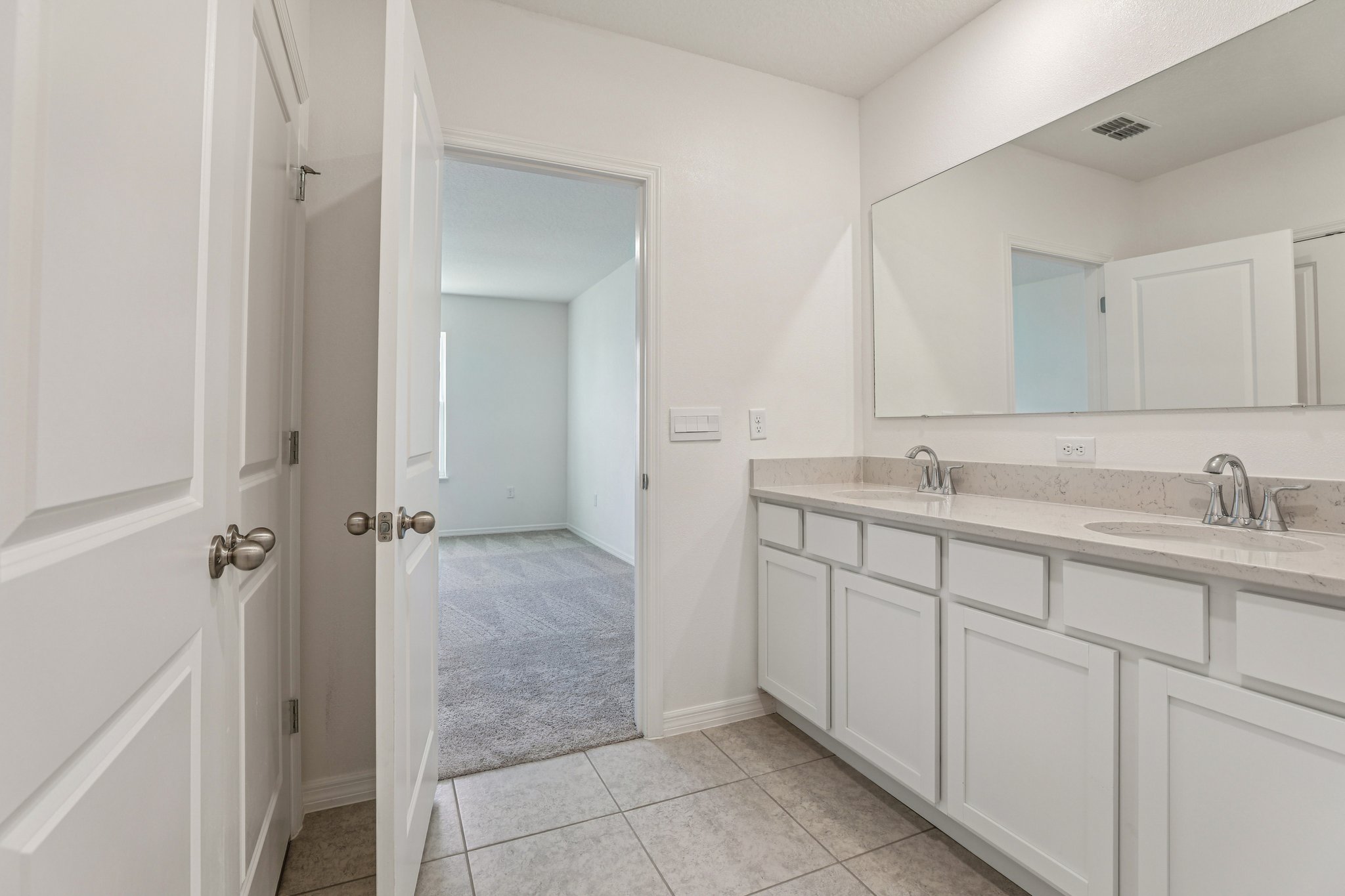 Spacious modern bathroom with a double sink vanity, large mirror, and tiled floor, leading to a carpeted bedroom.