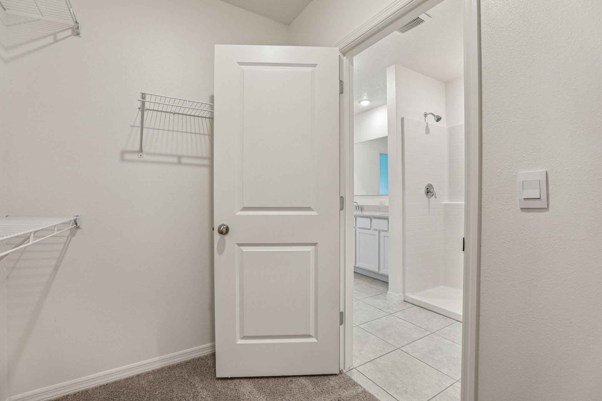 A spacious white closet with wire shelving leads into a bright bathroom featuring a shower and sink.