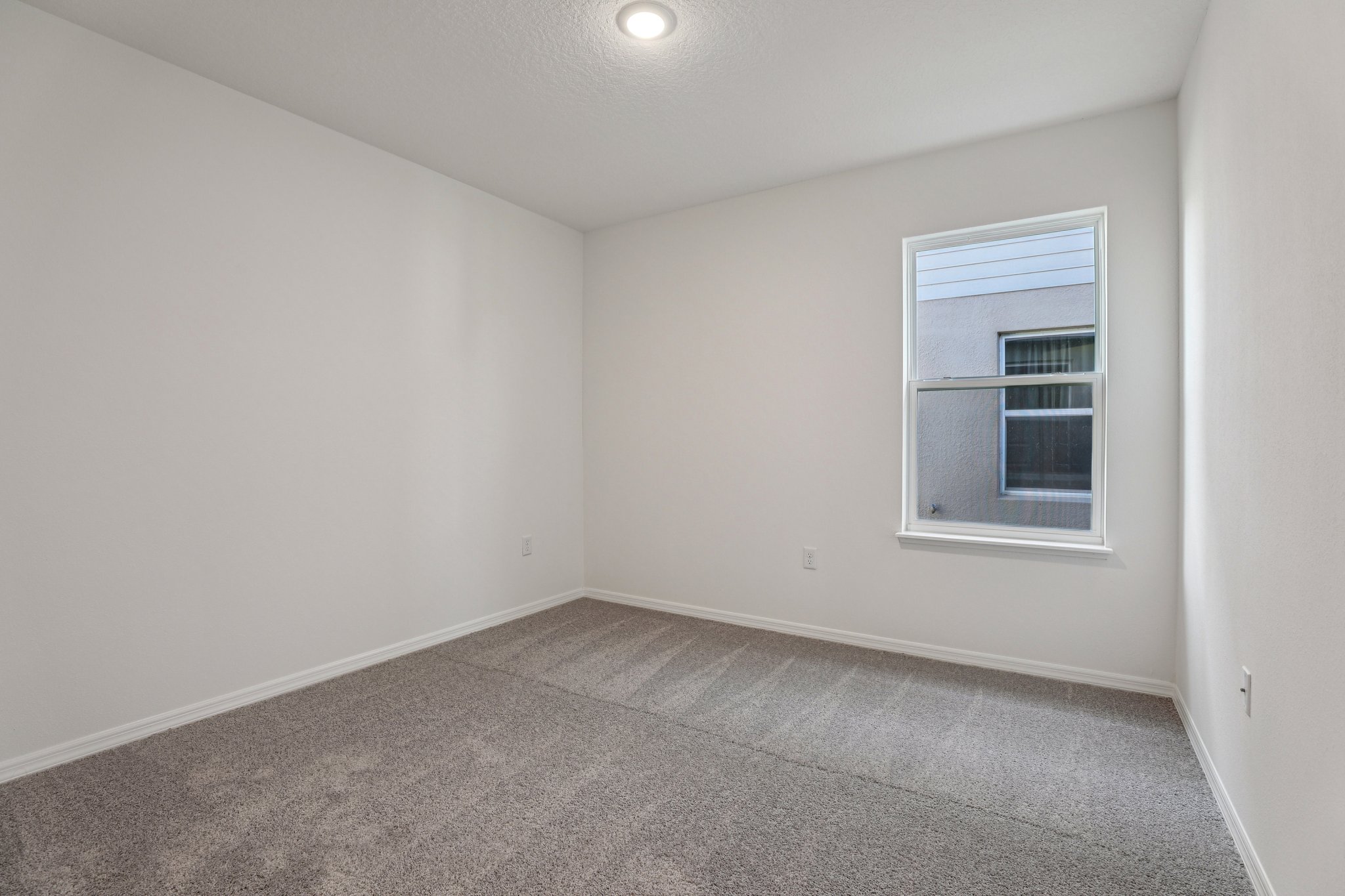 Minimalist empty room with gray carpet flooring, white walls, and a single window providing natural light.