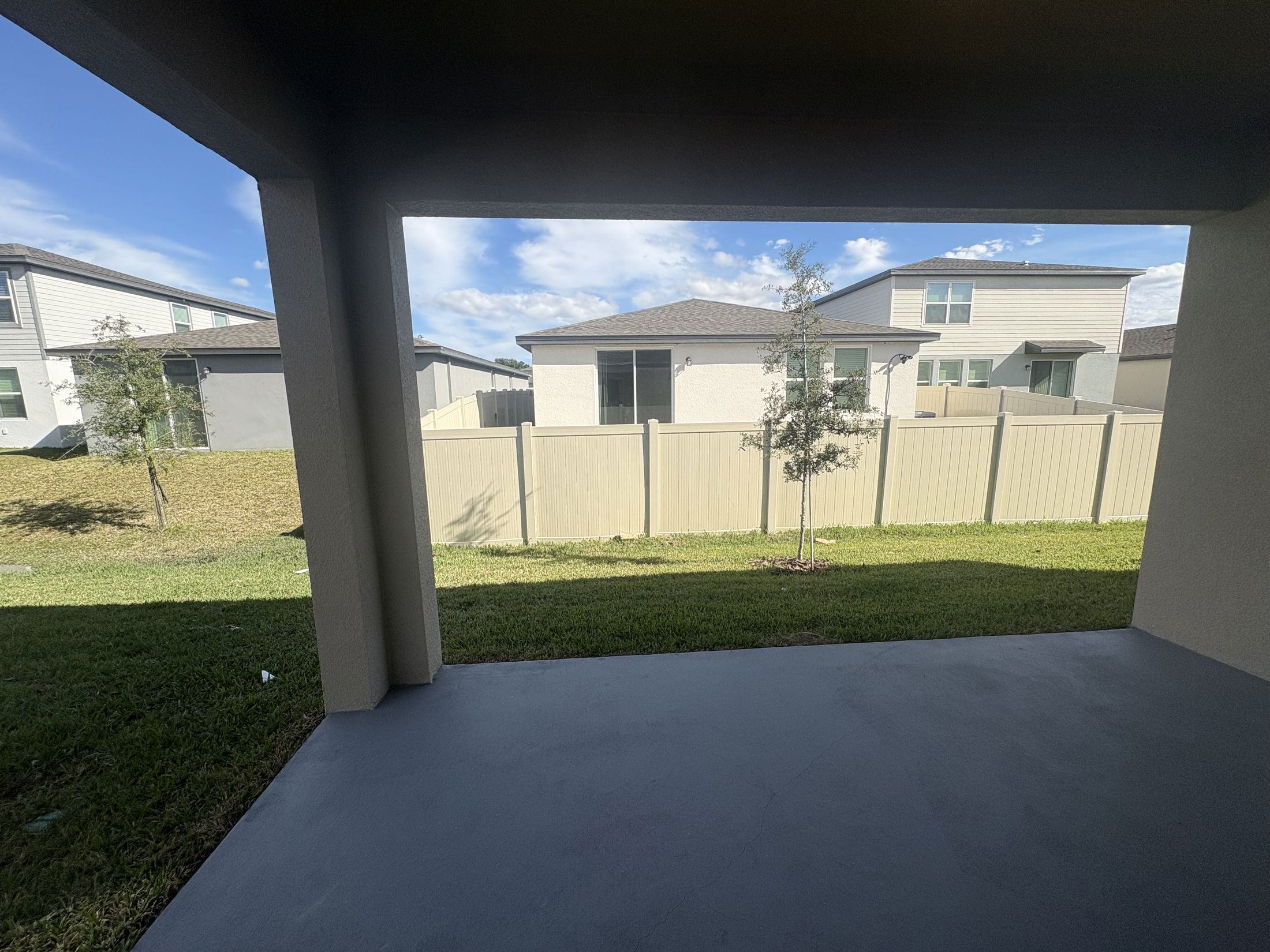 Covered patio view of suburban backyard with green lawn, white fence, and neighboring houses under a blue sky.