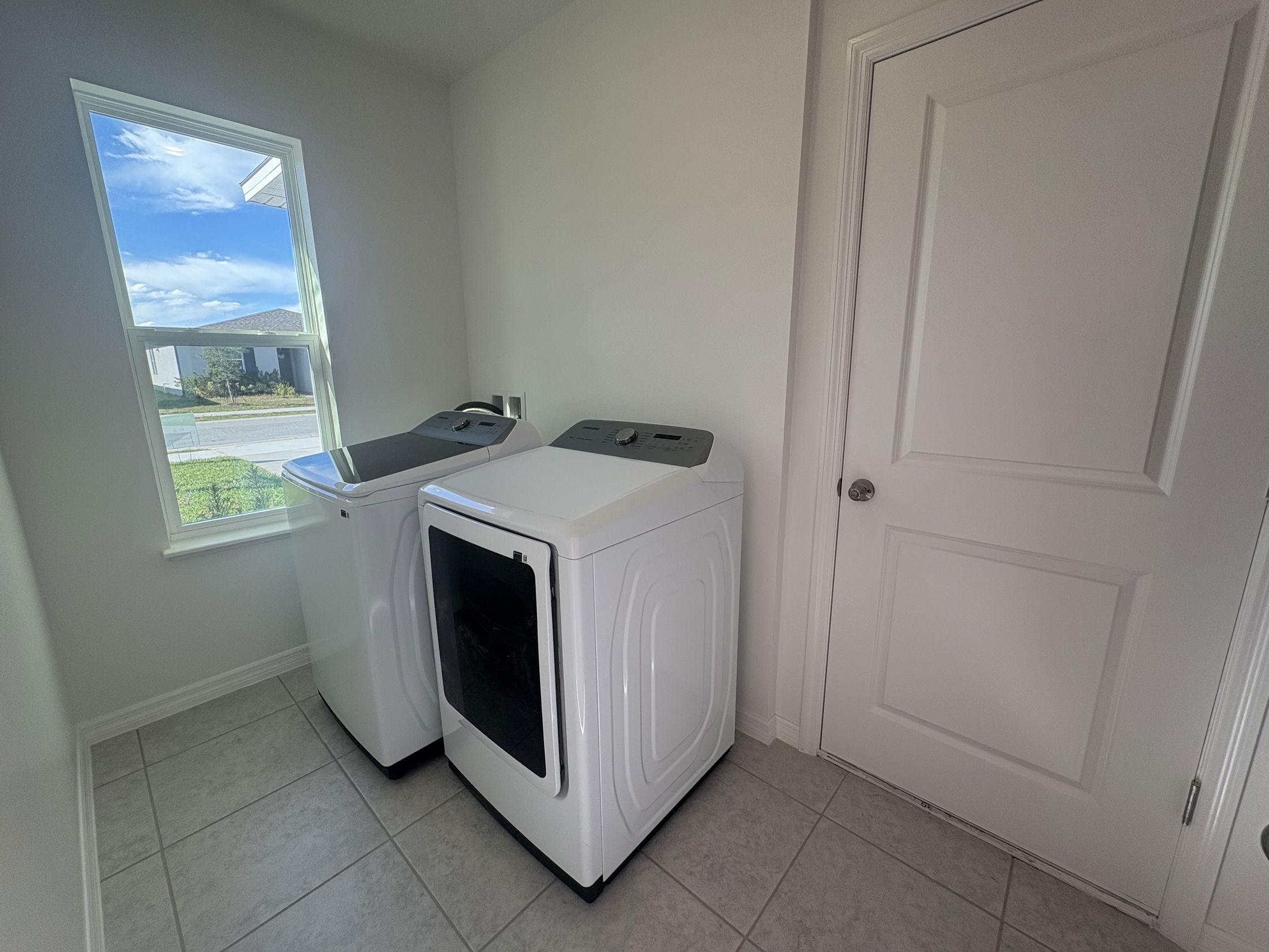 Bright laundry room with a top-loading washer, front-loading dryer, and a window overlooking a sunny neighborhood street.