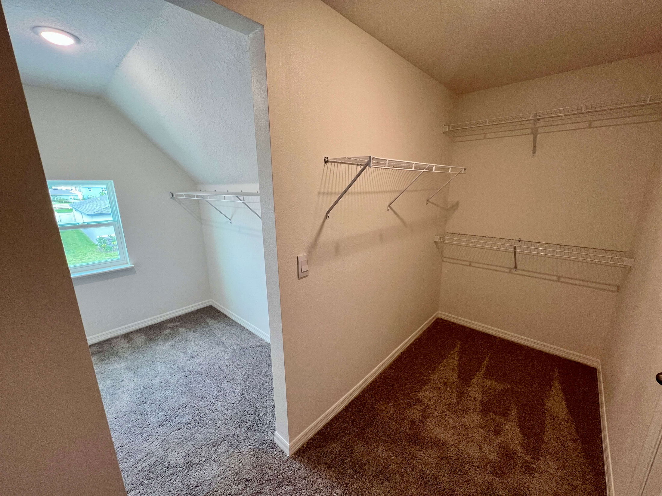 Spacious walk-in closet with carpeted floor, angled ceiling, and ample wire shelving, featuring a small window providing natural light.