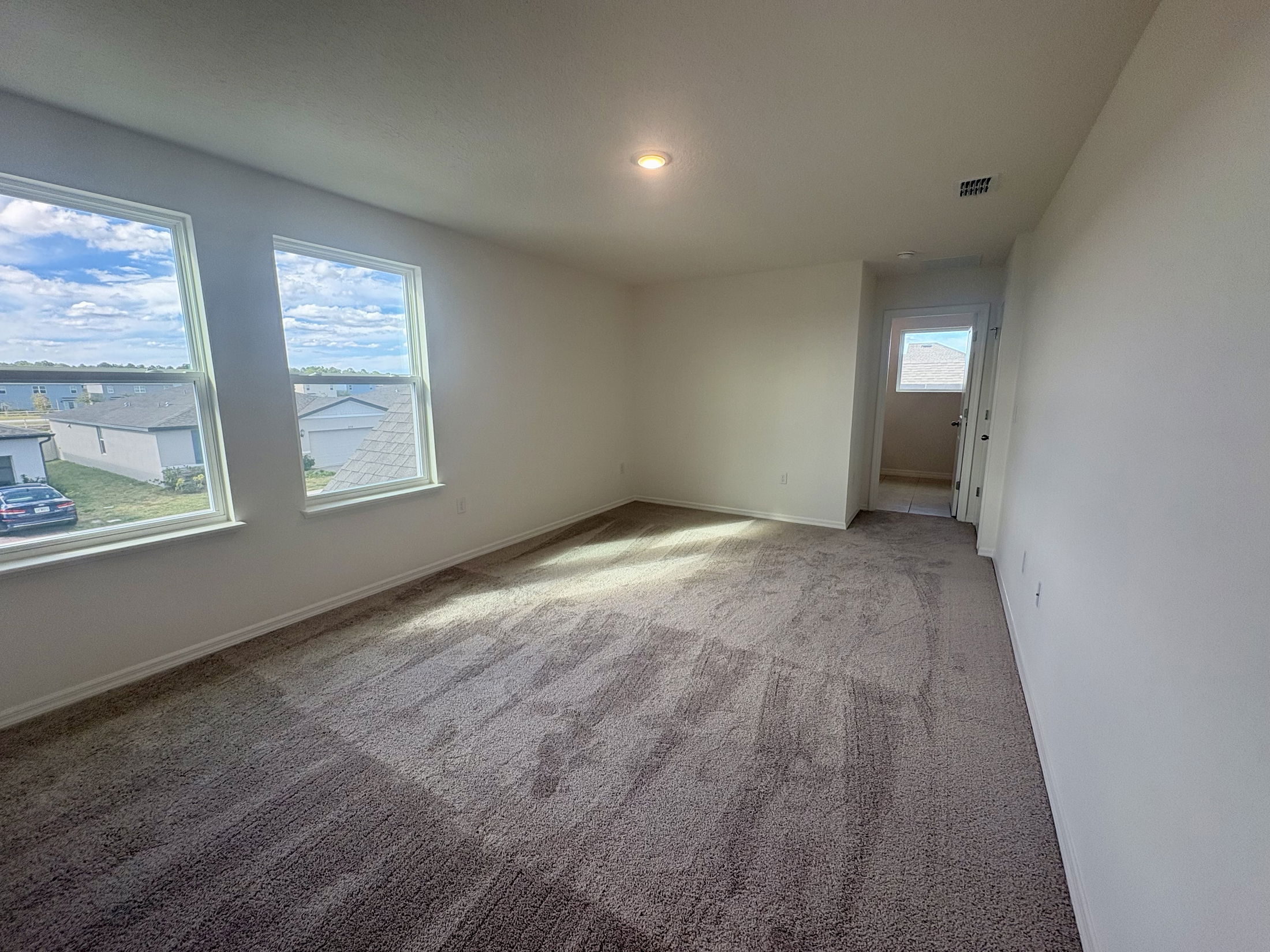 Bright, empty room with beige carpet, two large windows, and a view of suburban houses and sky.