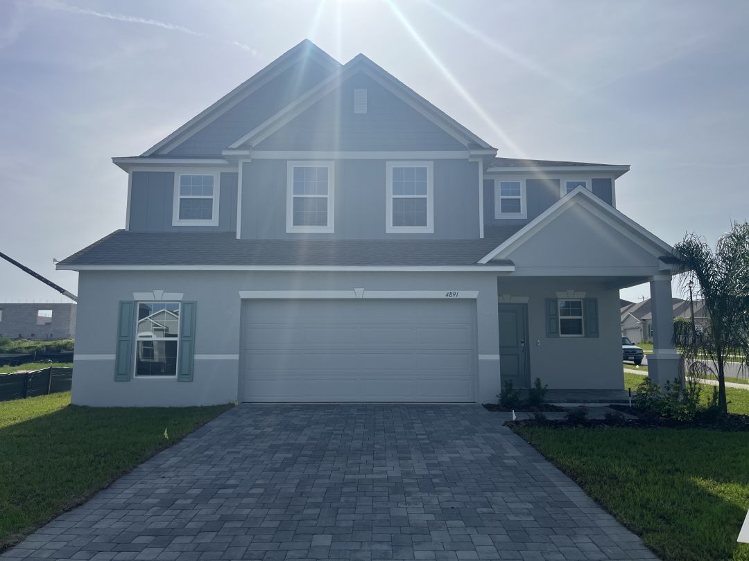 Two-story gray house with a gabled roof, double garage, and landscaped front yard, lit by midday sun.