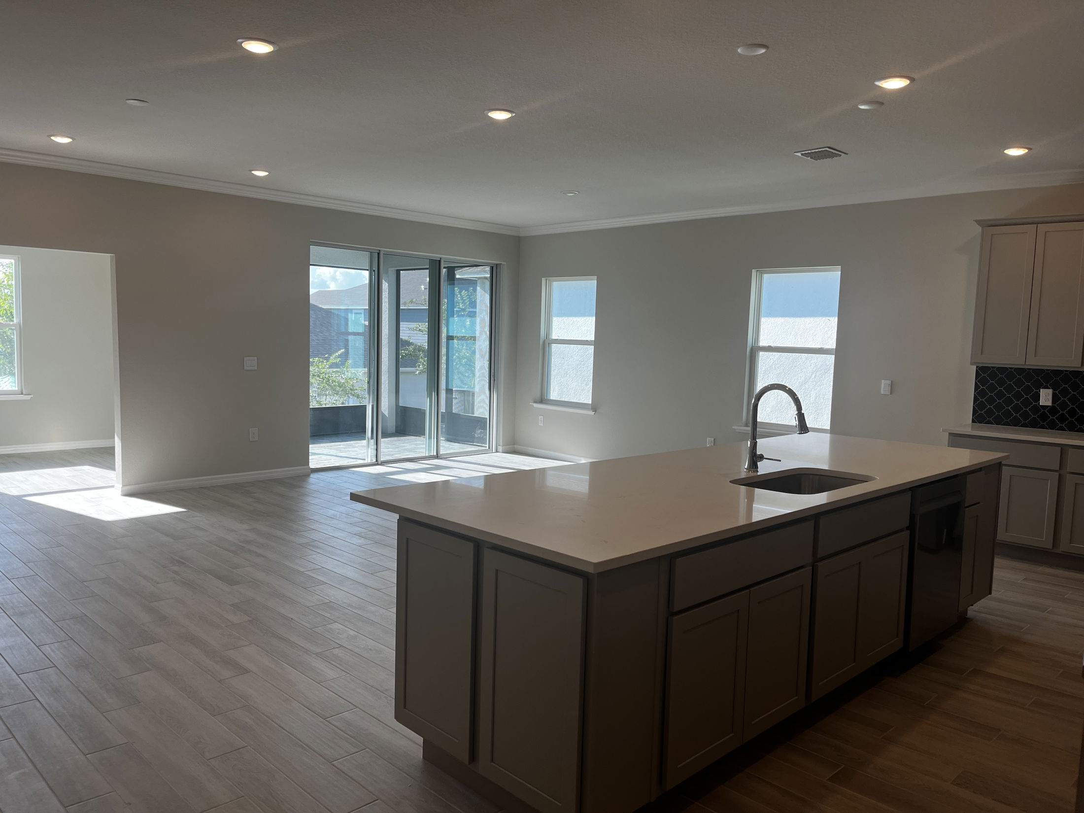 Modern kitchen and living area with wood flooring, an island sink, and large windows providing natural light.