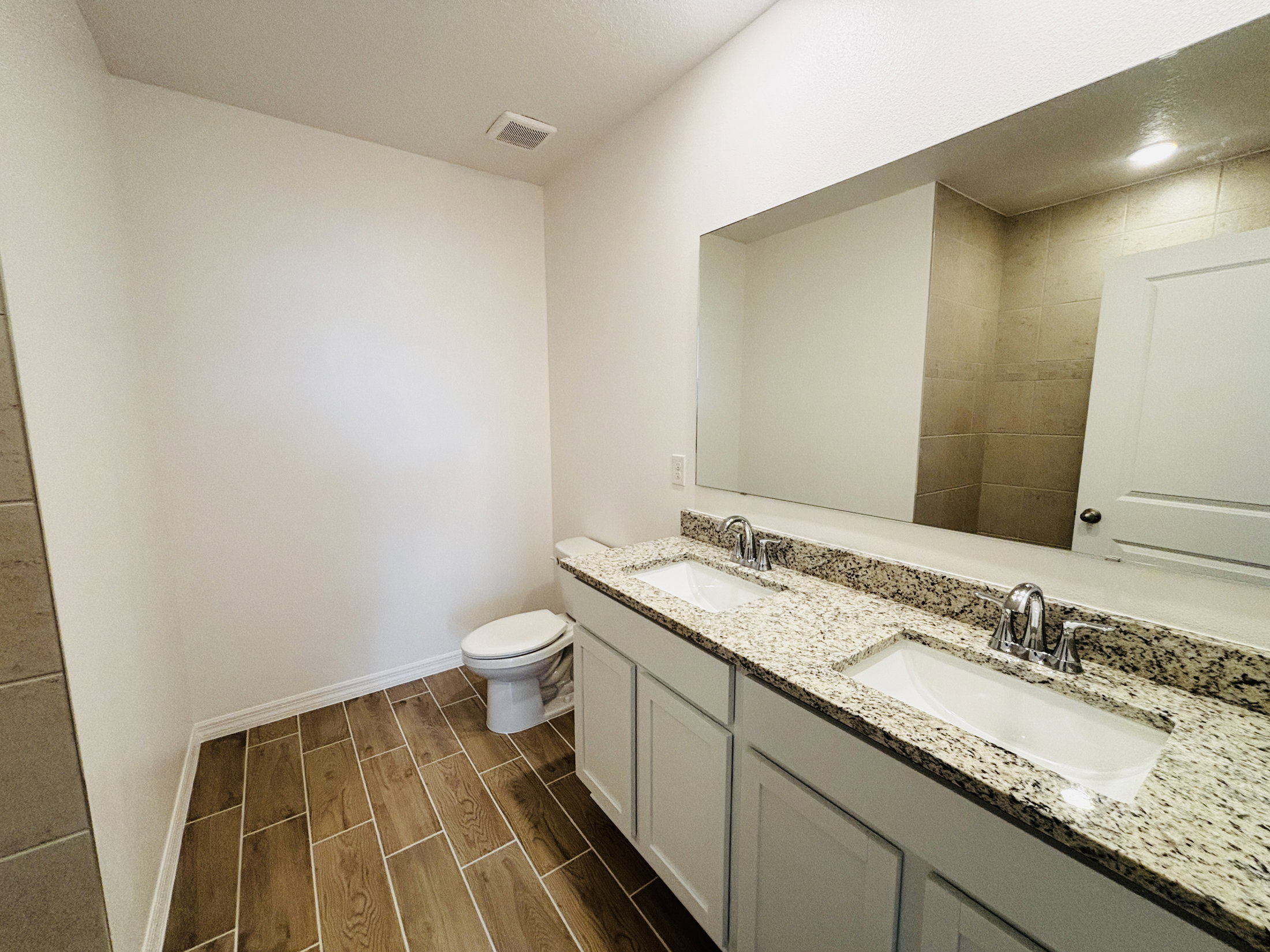 A modern bathroom featuring double sinks with a granite countertop, a large mirror, and a toilet in the corner.