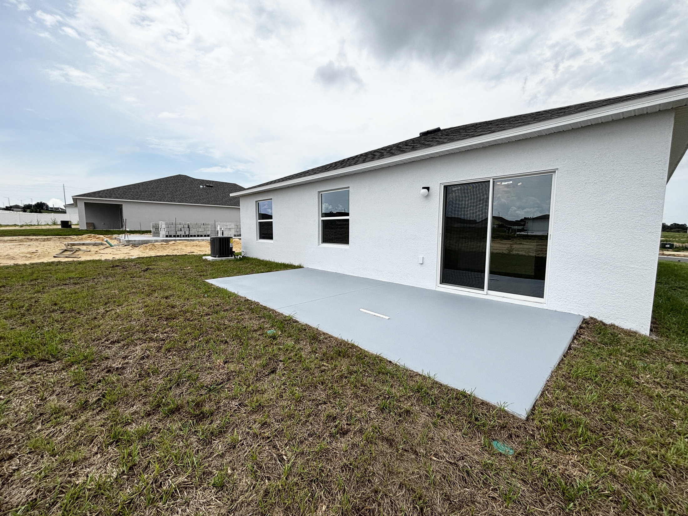 A white house with sliding glass doors and a concrete patio, surrounded by grassy land and a cloudy sky.