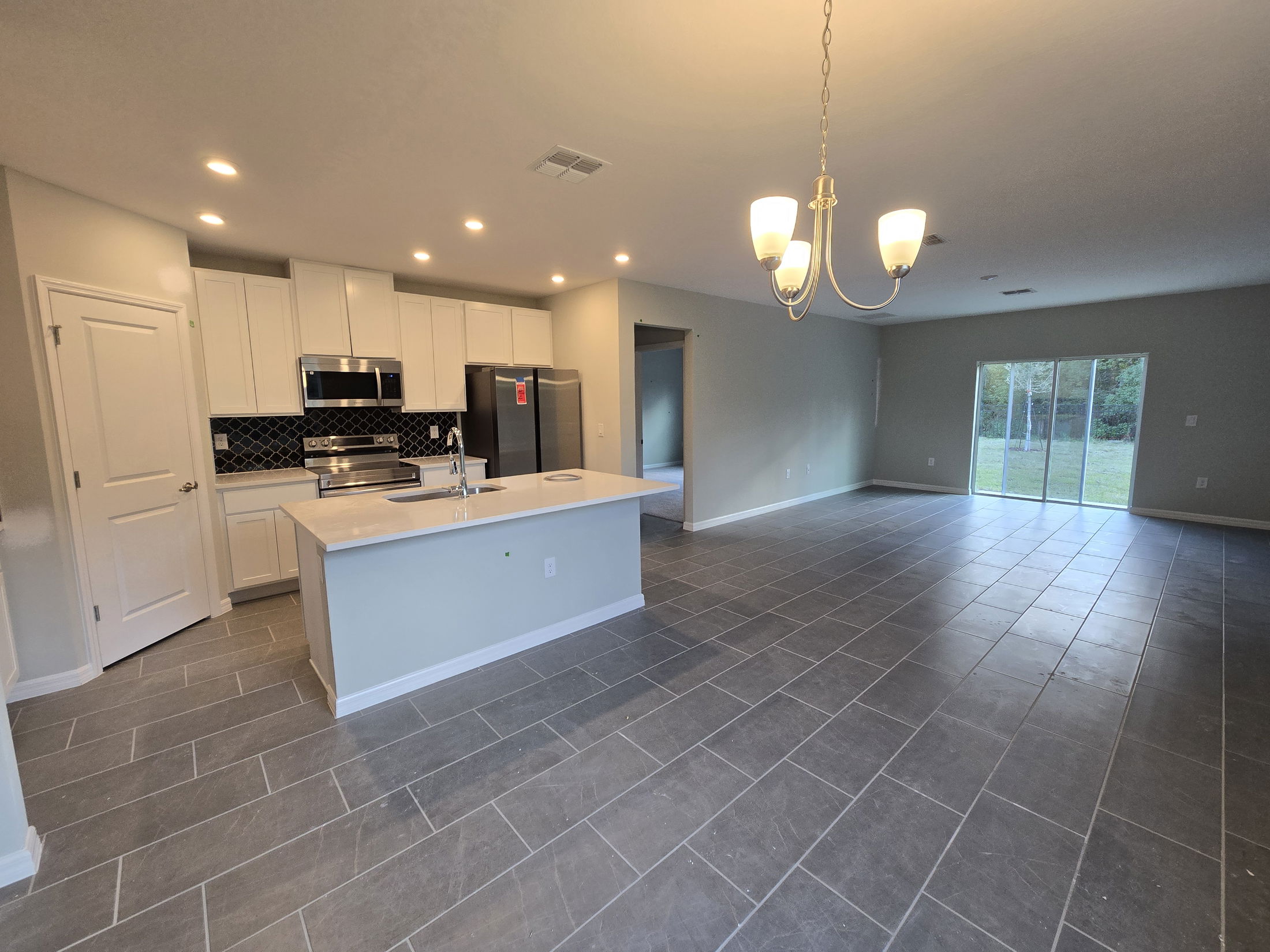 Modern open-concept kitchen and living area with tiled flooring, white cabinetry, stainless steel appliances, and a sliding glass door leading to a backyard.