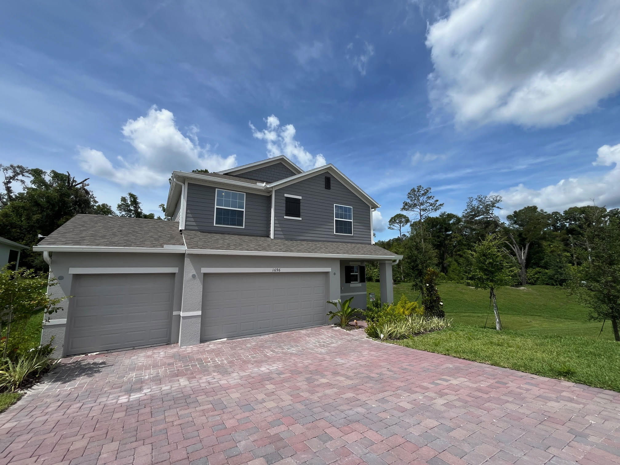Two-story modern gray house with a three-car garage, set against a lush green landscape and clear blue sky.