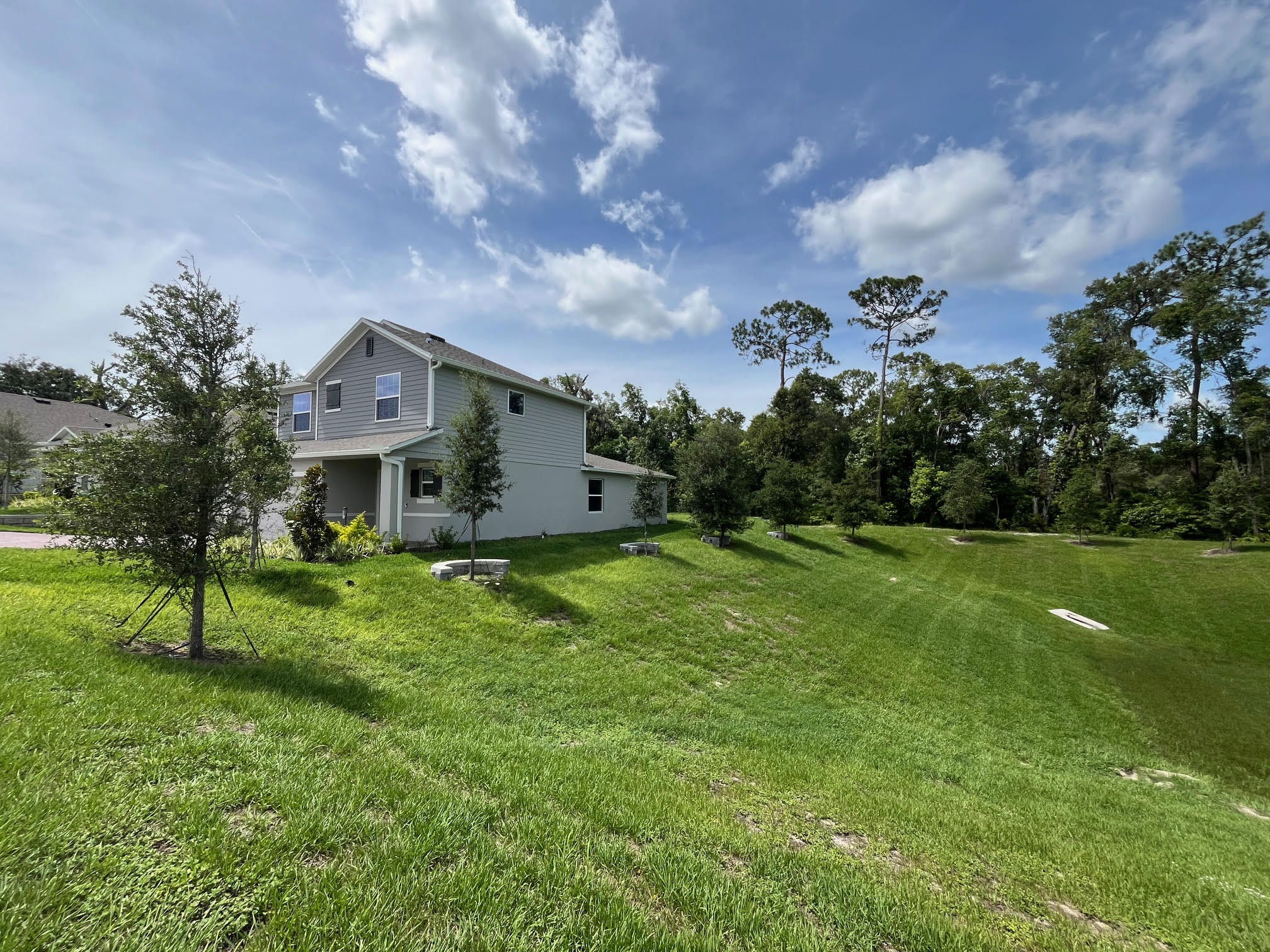 Modern two-story gray house with manicured lawn and trees, set against a backdrop of blue sky and clouds.