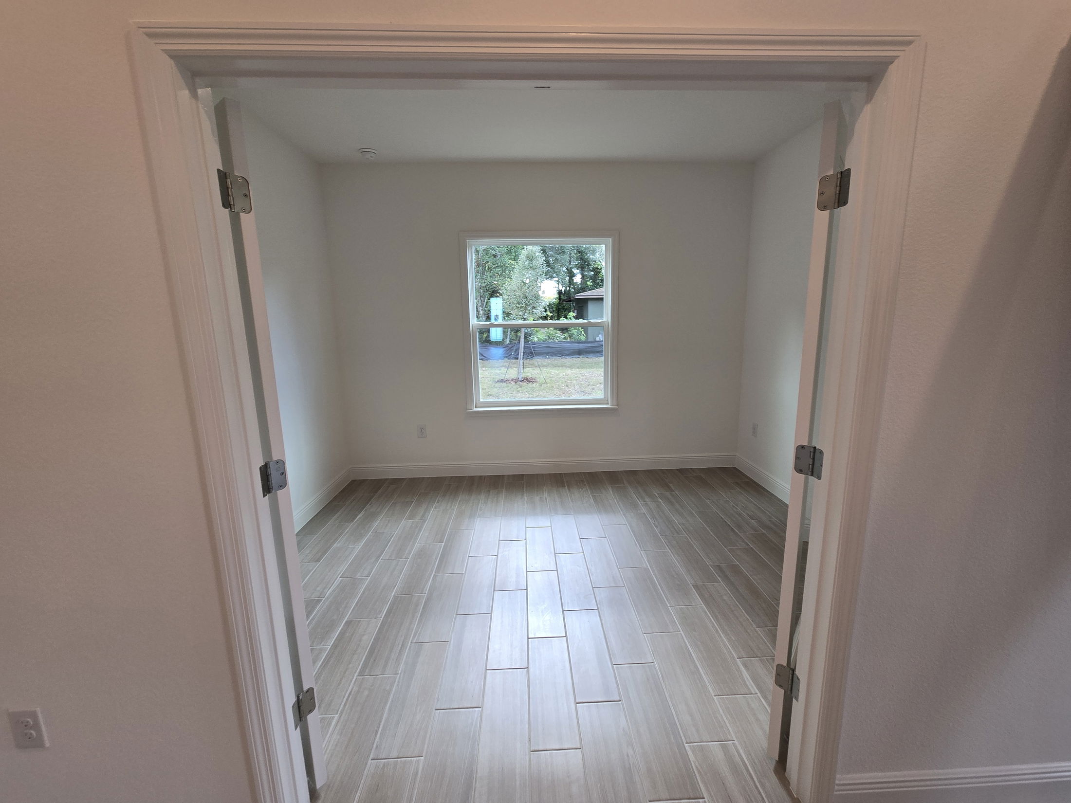 Empty room with wooden floor, white walls, and large window, viewed through open double doors.