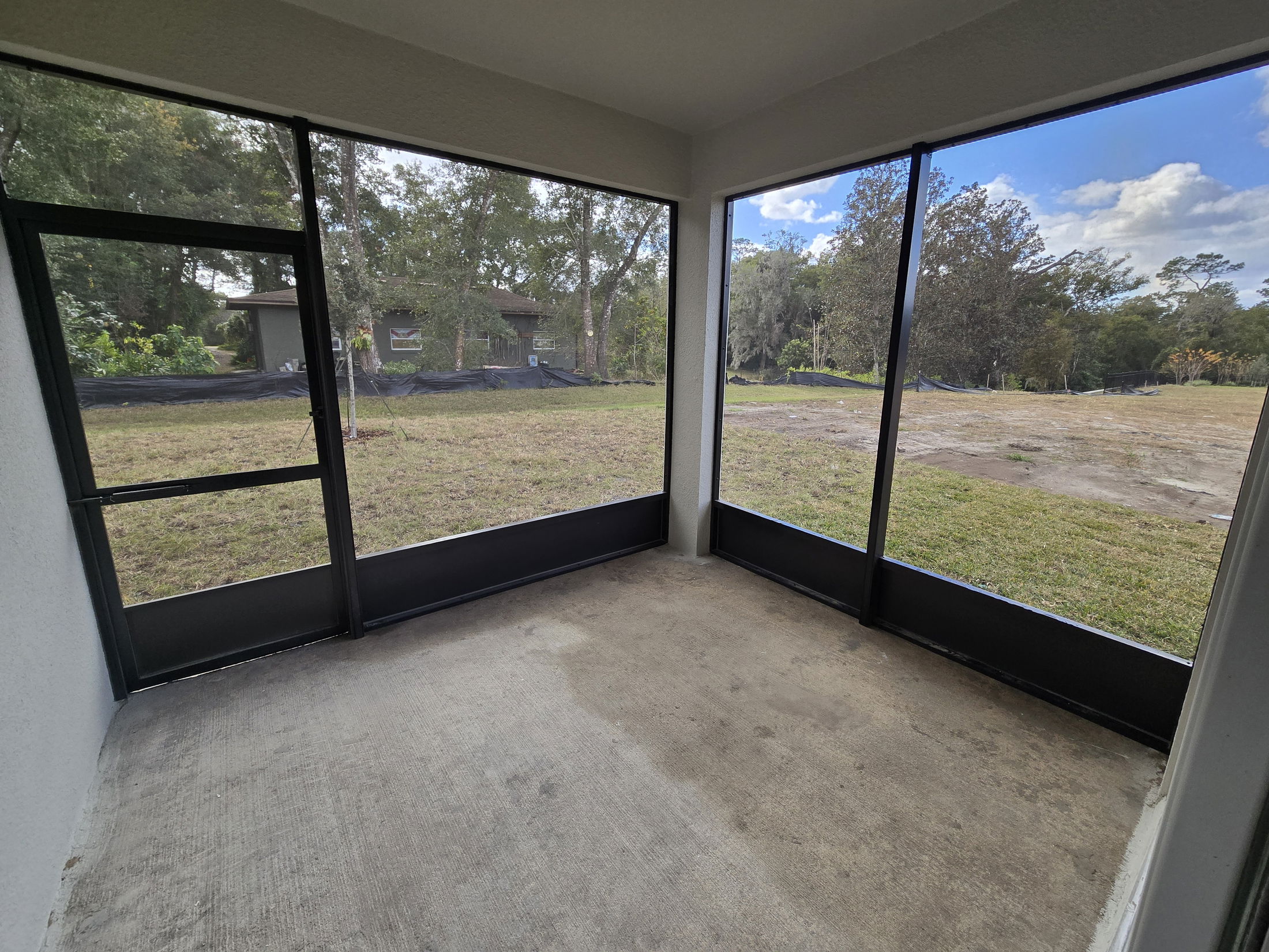 Enclosed patio with a screen wall and concrete floor overlooking a grassy yard with trees under a blue sky.