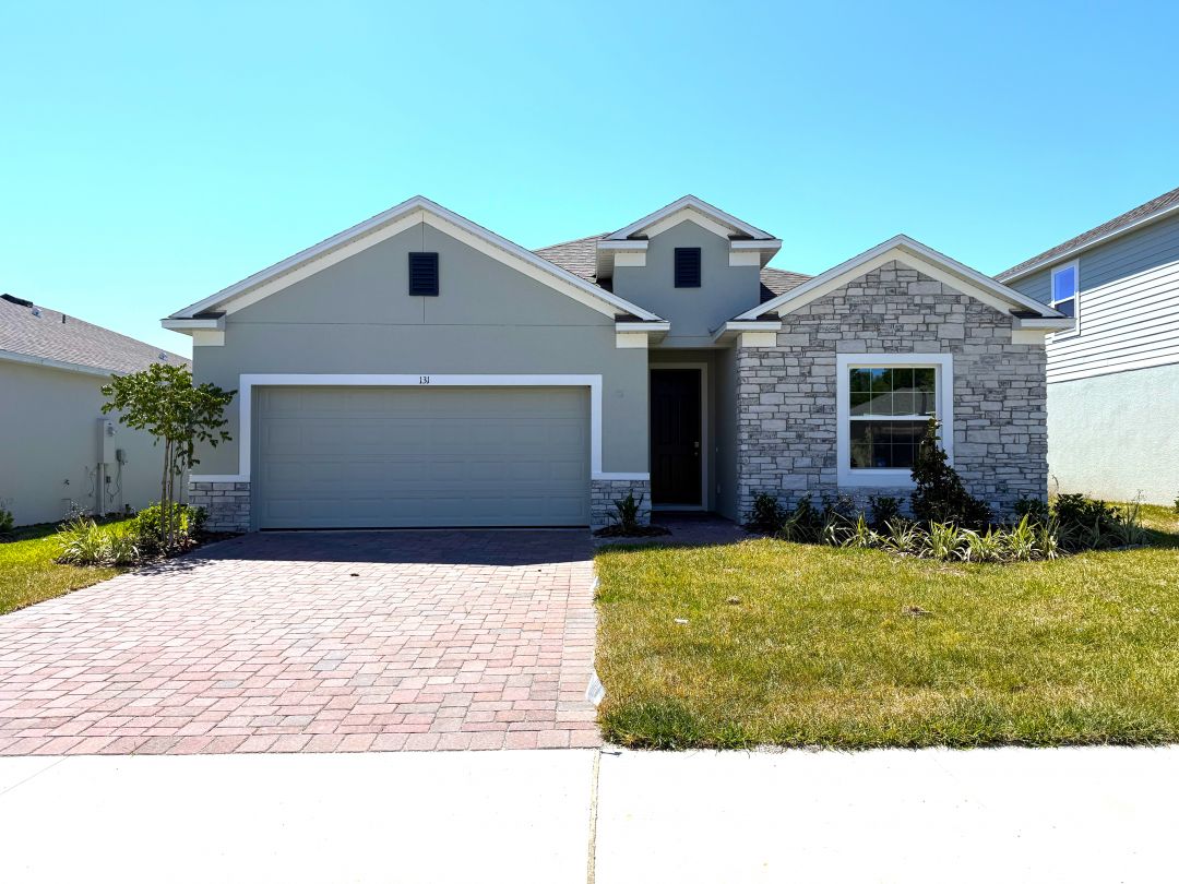 Modern single-story home with a two-car garage, brick driveway, and well-maintained front yard featuring stone accents and a clear blue sky backdrop.