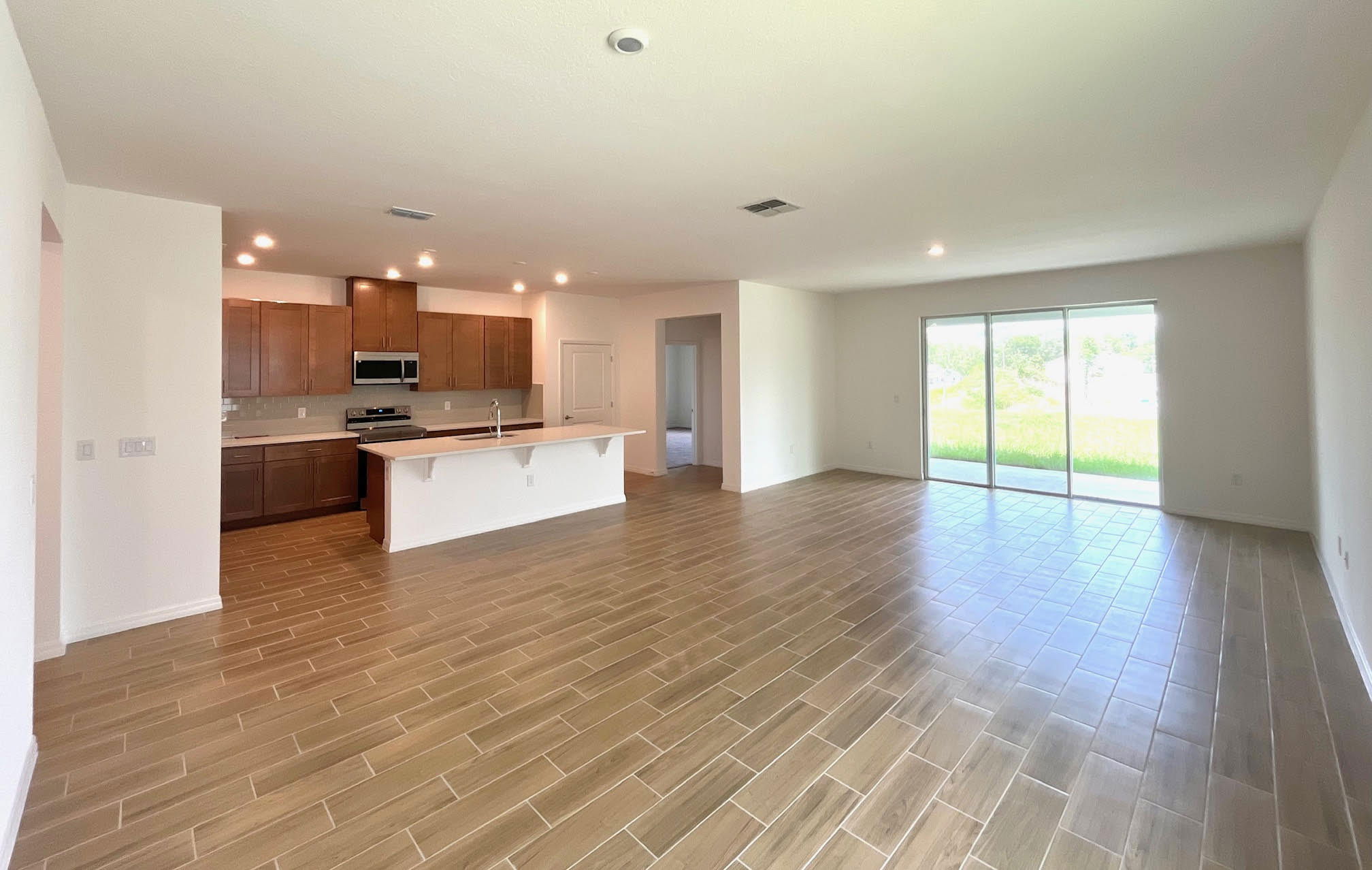 Spacious open-plan living area with modern kitchen, wooden tile flooring, and large sliding glass doors leading to a backyard.