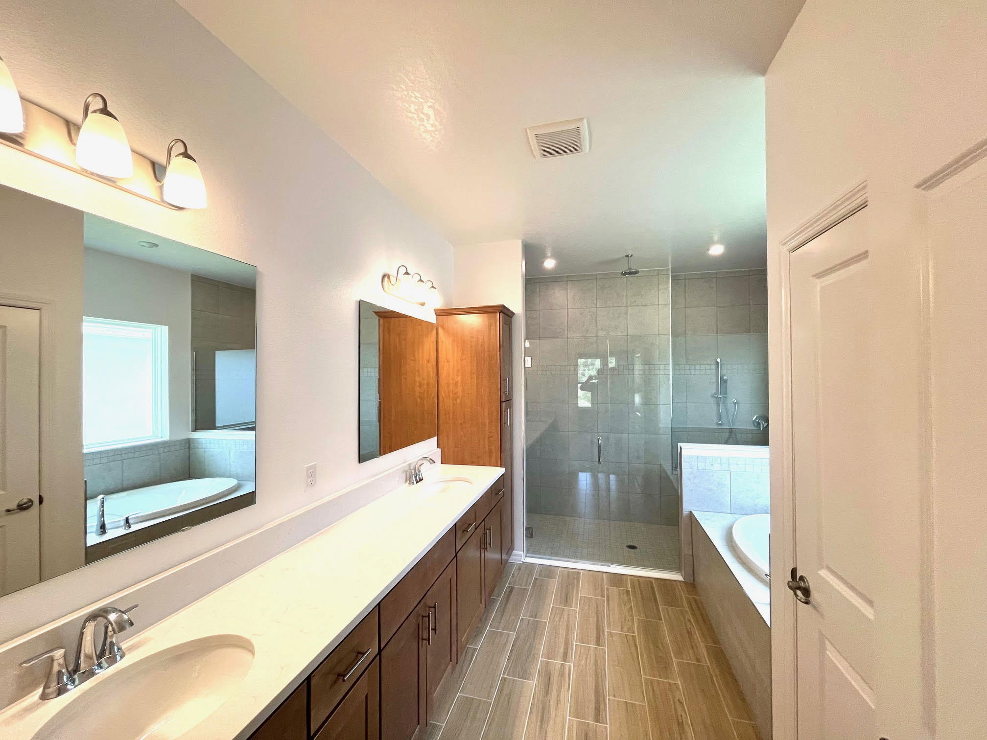 Modern bathroom interior featuring a double vanity with marble countertop, large glass-enclosed shower, bathtub, and wooden cabinets under bright overhead lighting.