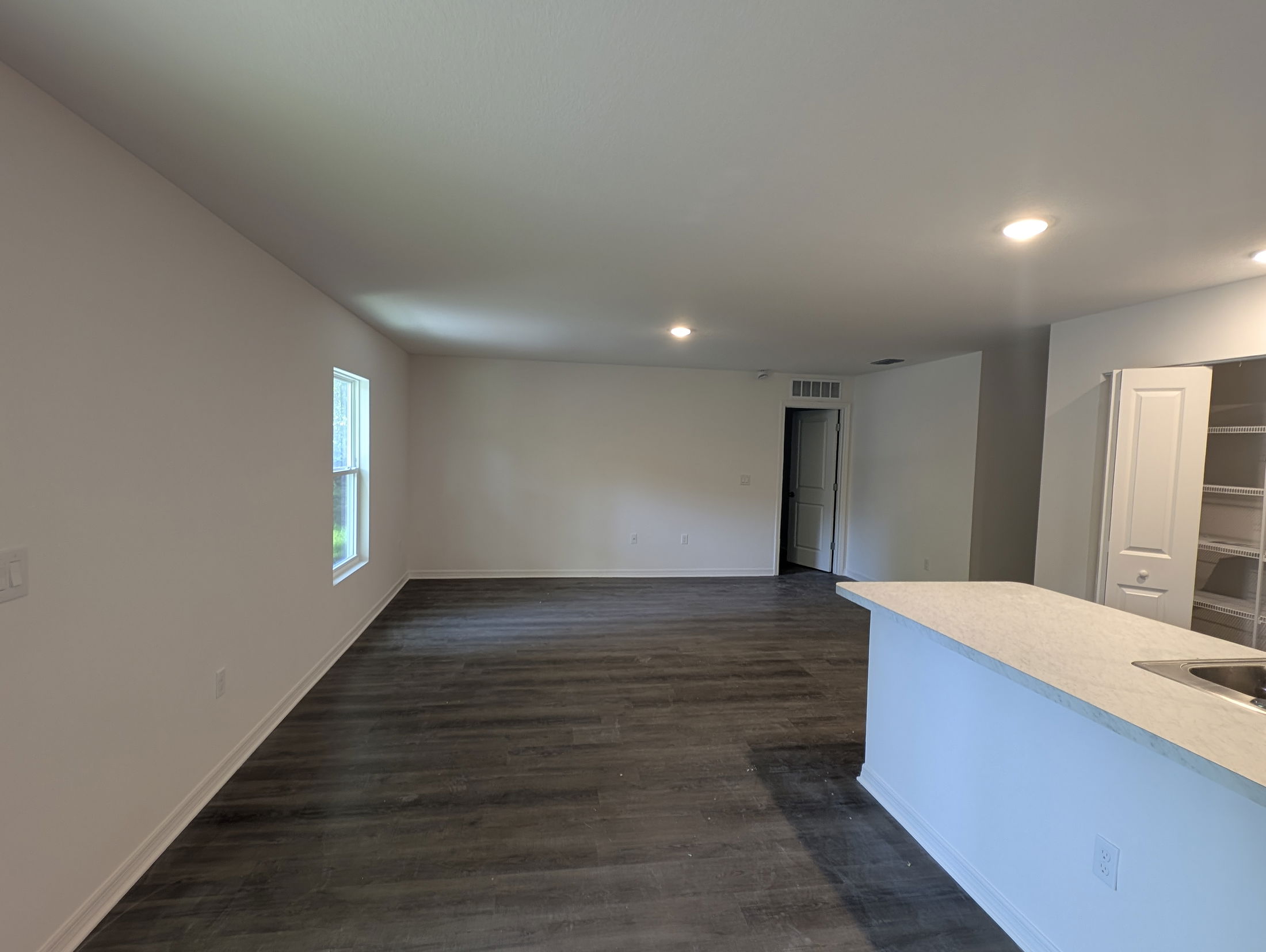 Bright, modern living space with dark wood flooring and white walls, featuring a kitchen peninsula and illuminated by recessed lighting.