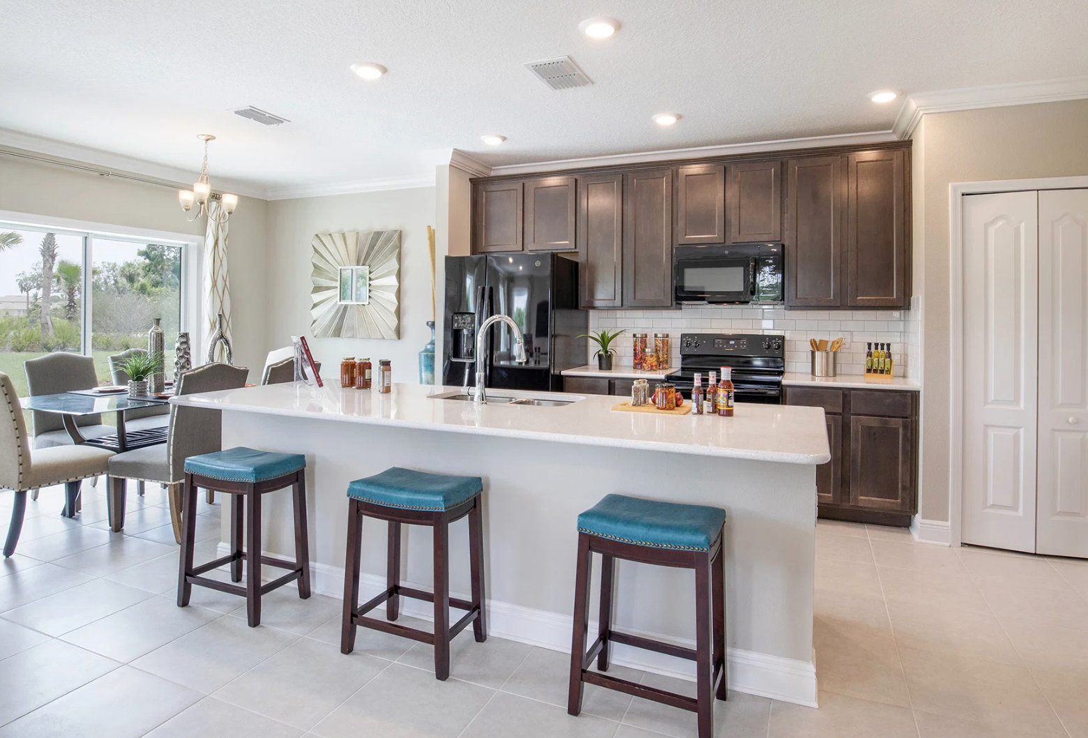Modern kitchen featuring dark wooden cabinets, a white countertop, and bar stools, with a dining area visible in the background.