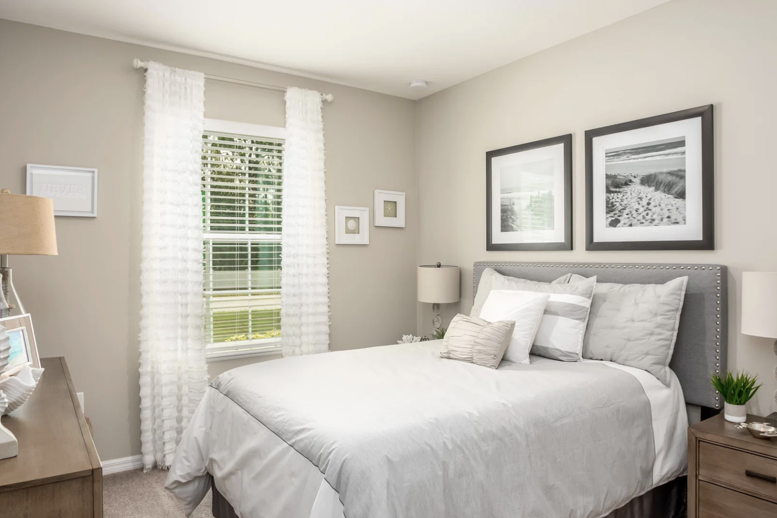 A cozy bedroom featuring a gray upholstered bed, white bedding, framed photographs on the wall, and a window with sheer curtains.